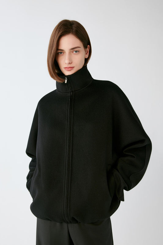 A model wearing a black zip-up jacket with a high mock neck collar, photographed against a neutral light gray background.