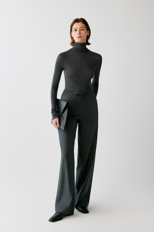 A model stands against a light gray studio background wearing a dark charcoal gray ribbed turtleneck sweater and matching wide-leg trousers, holding a dark clutch.