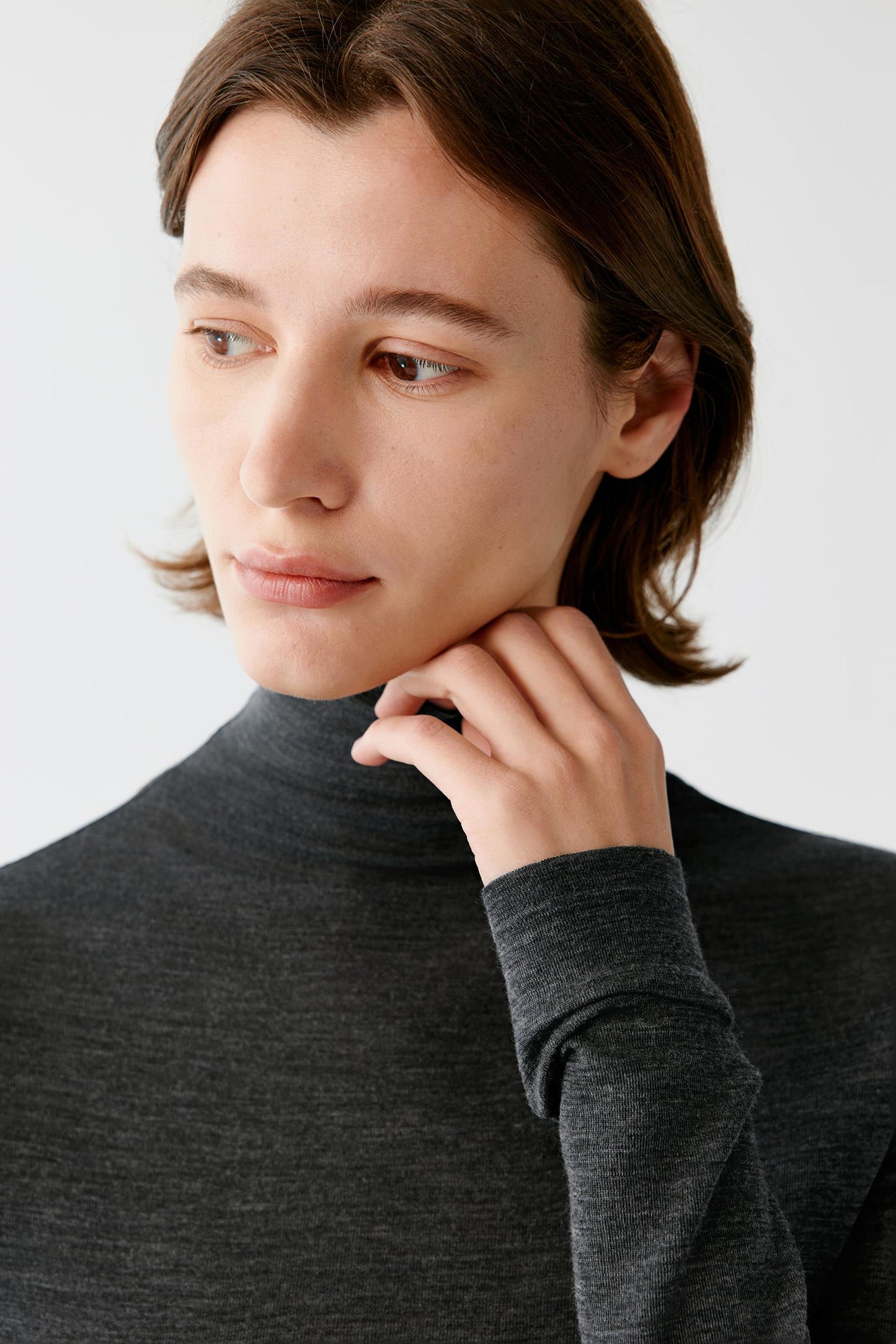 A close-up of a person wearing a dark charcoal gray ribbed turtleneck sweater against a light gray background.