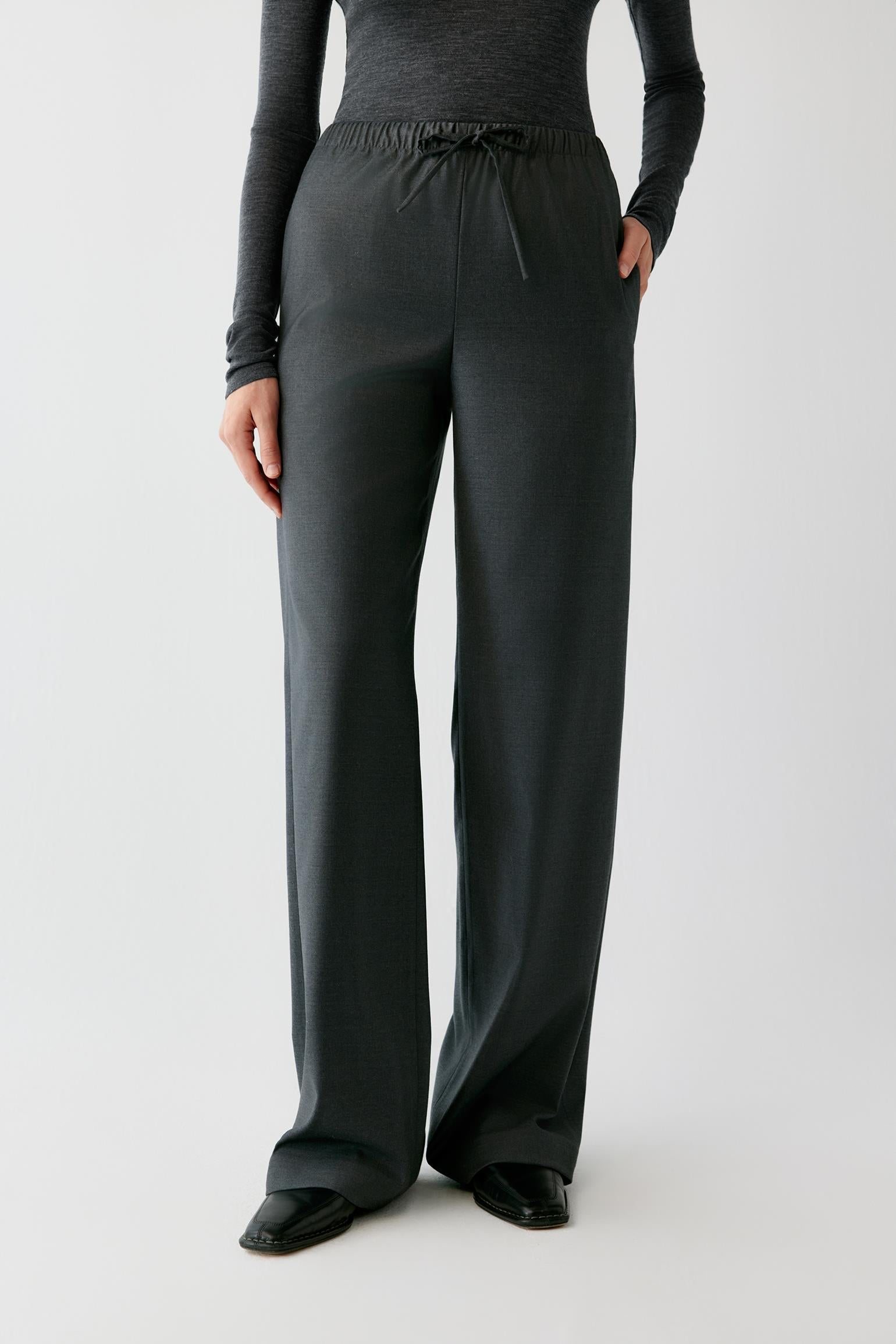 A model wearing dark charcoal gray wide-leg lounge pants with an elastic drawstring waist, paired with a matching dark gray long-sleeve top and black shoes, against a light gray studio background.