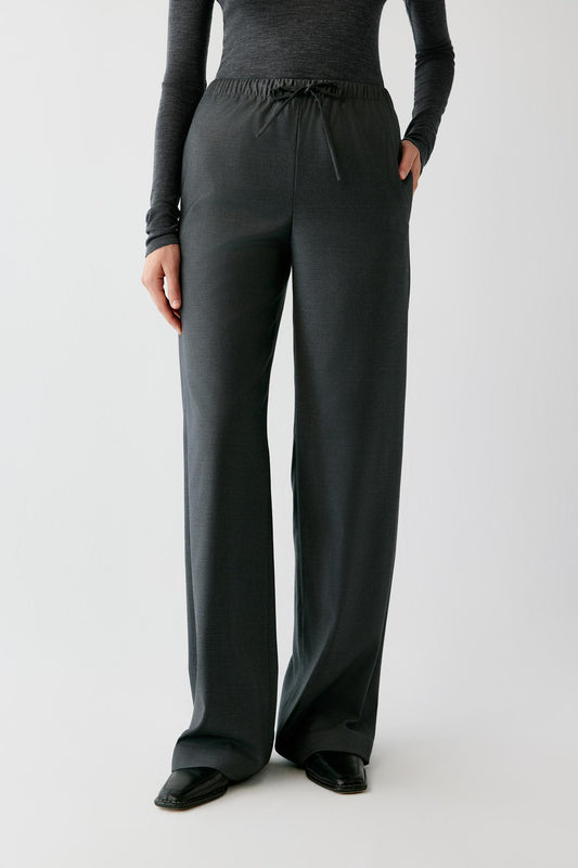 A model wearing dark charcoal gray wide-leg lounge pants with an elastic drawstring waist, paired with a matching dark gray long-sleeve top and black shoes, against a light gray studio background.