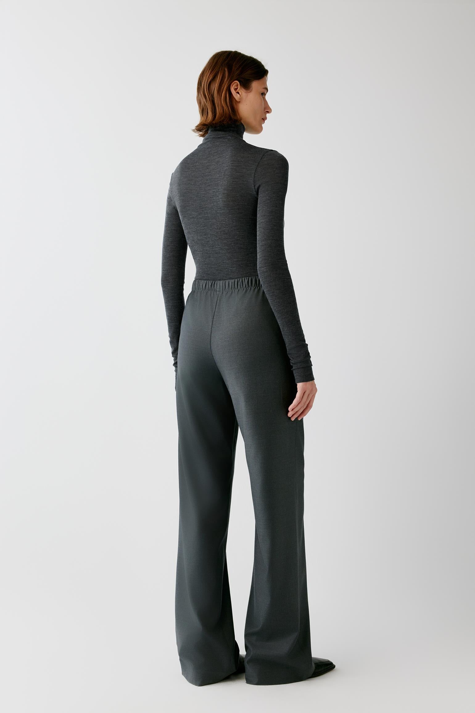 A woman shown from the back wearing a dark gray ribbed knit mock-neck long-sleeve top and matching wide-leg trousers, photographed against a light gray studio background.