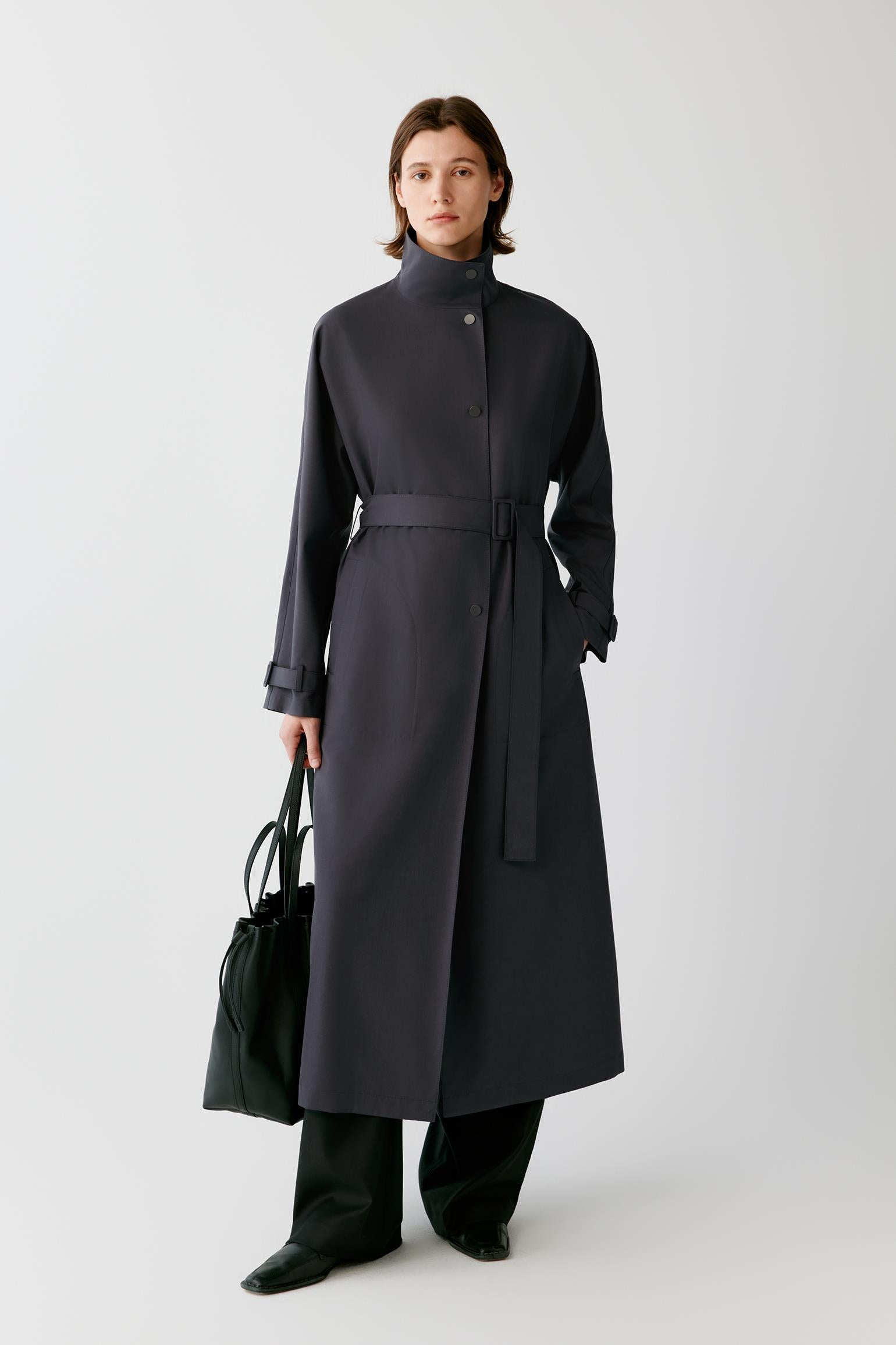 A model stands against a light gray studio background wearing a long dark charcoal-gray belted coat with a high collar, paired with dark trousers and holding a black tote bag.