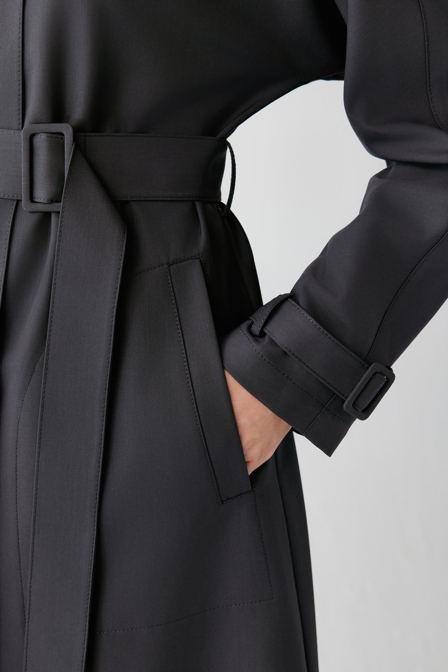 Close-up of a black trench coat with a matching waist belt, shown on a model with one hand in the pocket. The coat features structured shoulders, a belt buckle detail, and a clean light gray studio background.