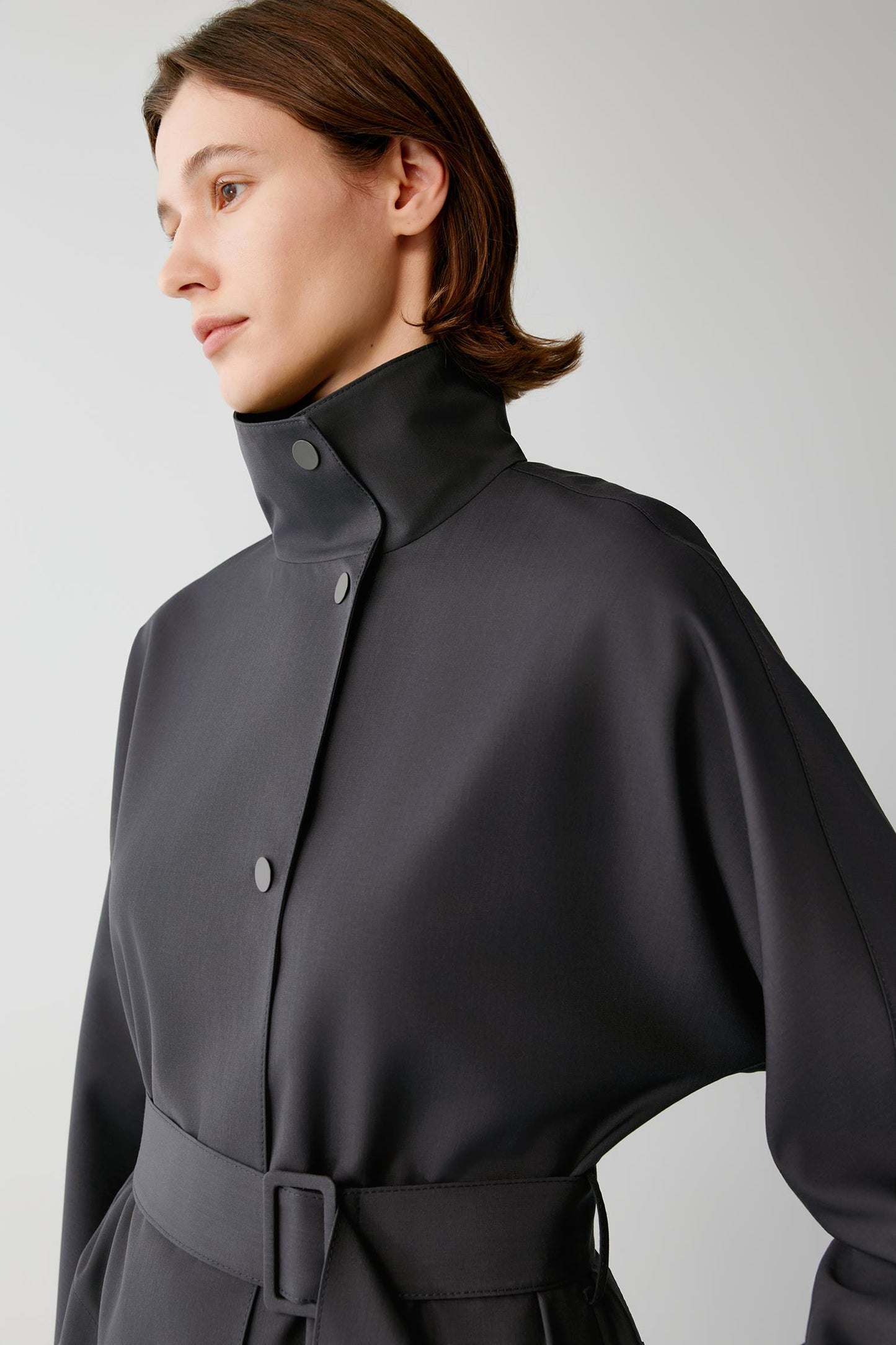 A close-up of a model wearing a dark gray high-neck, long-sleeve coat with a belted waist and snap-button front, set against a plain light gray background.