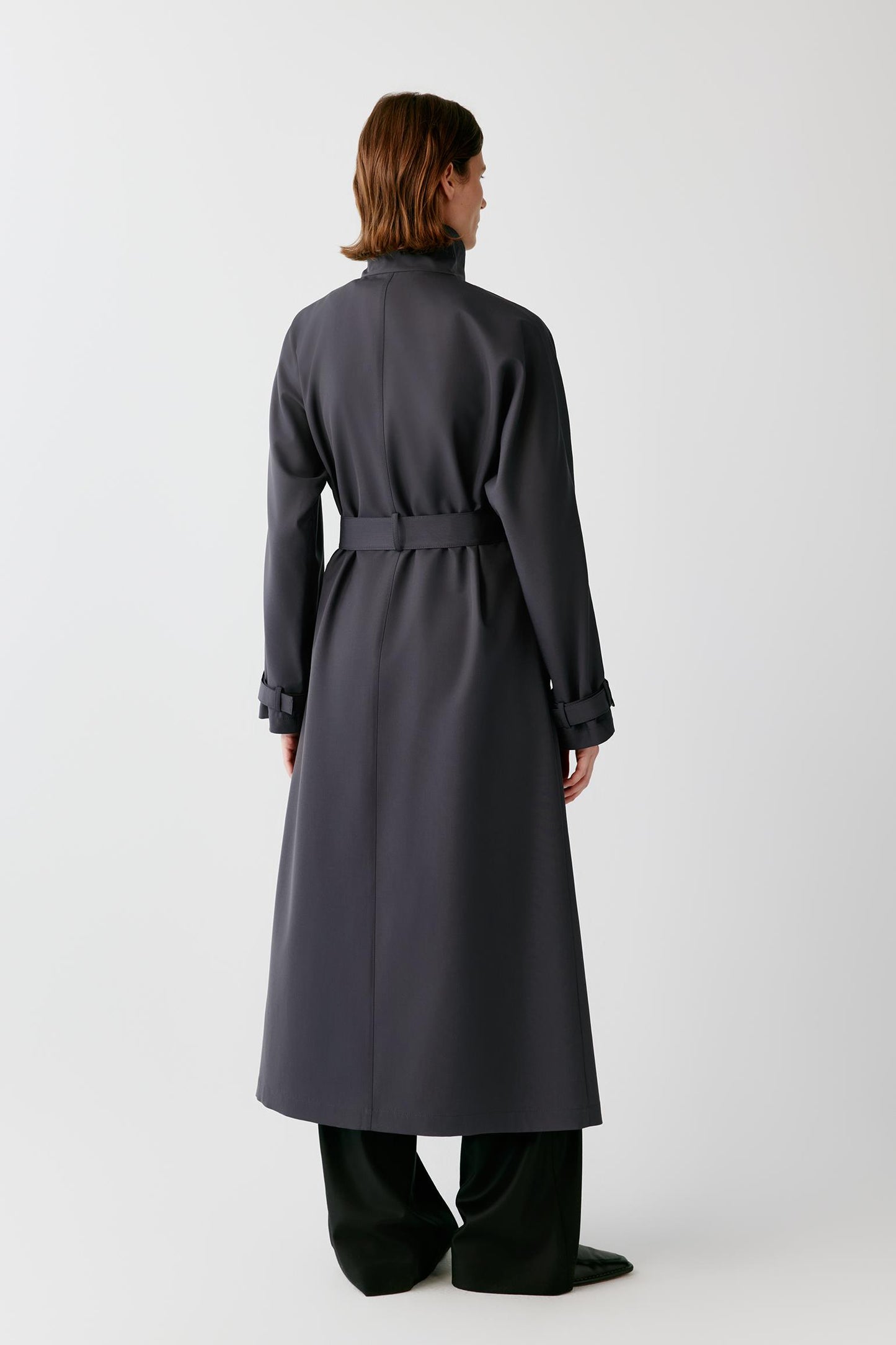A model viewed from the back wearing a dark charcoal gray long trench coat with a matching waist belt, paired with black wide-leg trousers, against a light gray studio background.