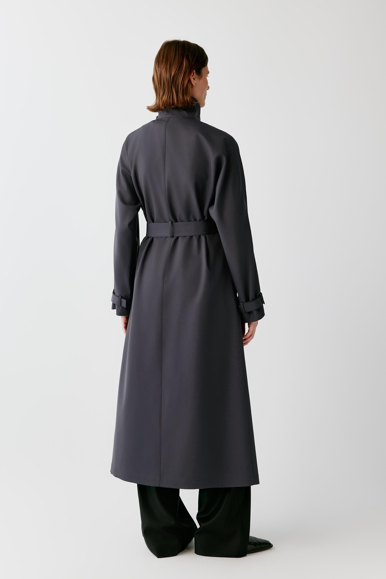 A model viewed from the back wearing a dark charcoal gray long trench coat with a matching waist belt, paired with black wide-leg trousers, against a light gray studio background.