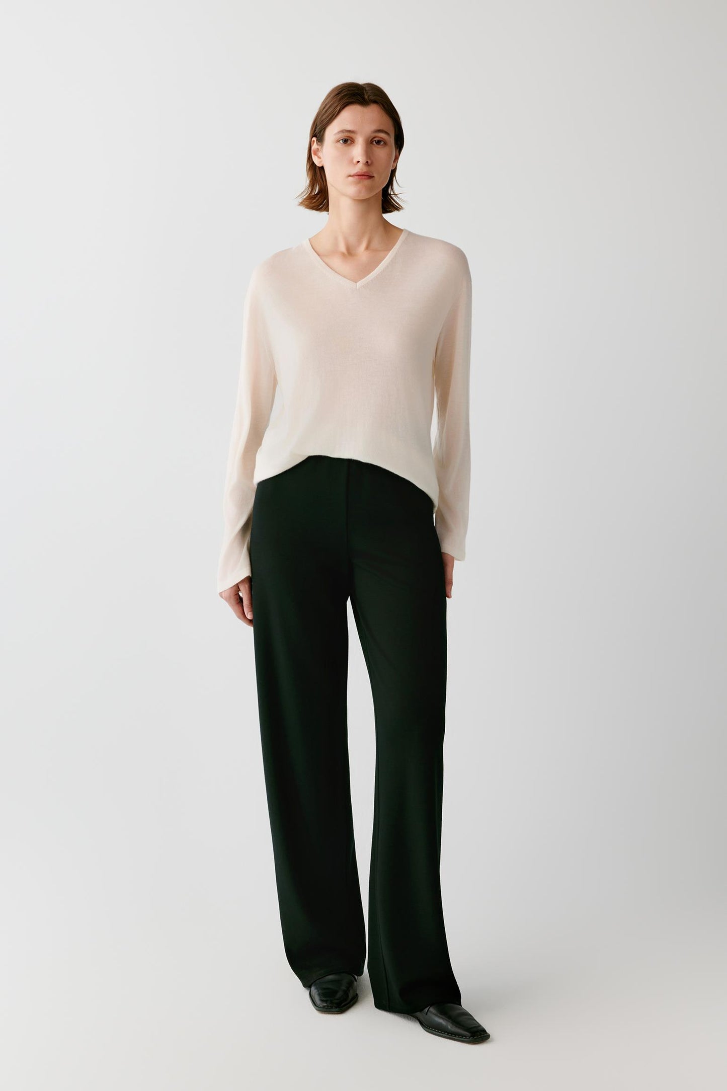 A model stands against a light gray studio background wearing a cream V-neck knit sweater and dark green wide-leg trousers.