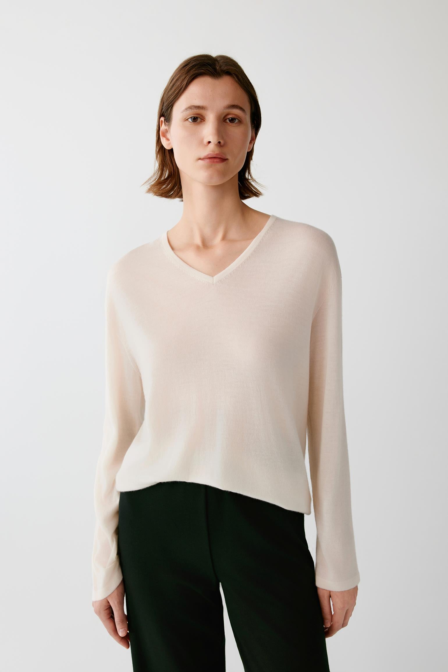 A woman modeling a light beige long-sleeve V-neck knit sweater with dark green trousers, photographed against a plain light gray studio background.