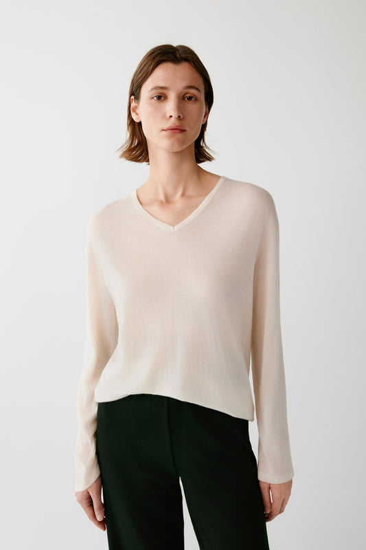 A woman modeling a light beige long-sleeve V-neck knit sweater with dark green trousers, photographed against a plain light gray studio background.