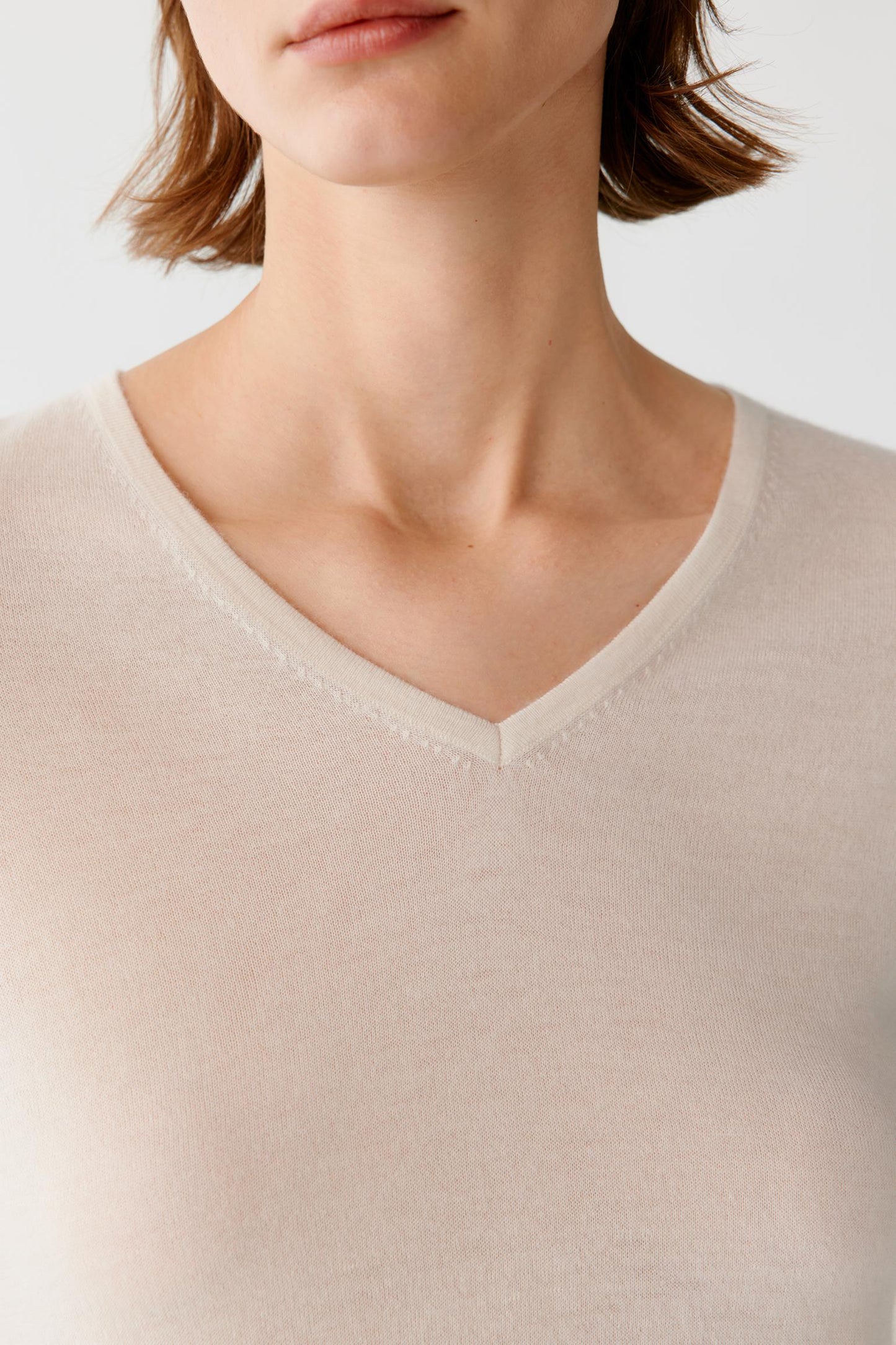 Close-up of a model wearing a light beige or cream V-neck knit top against a plain light gray background.