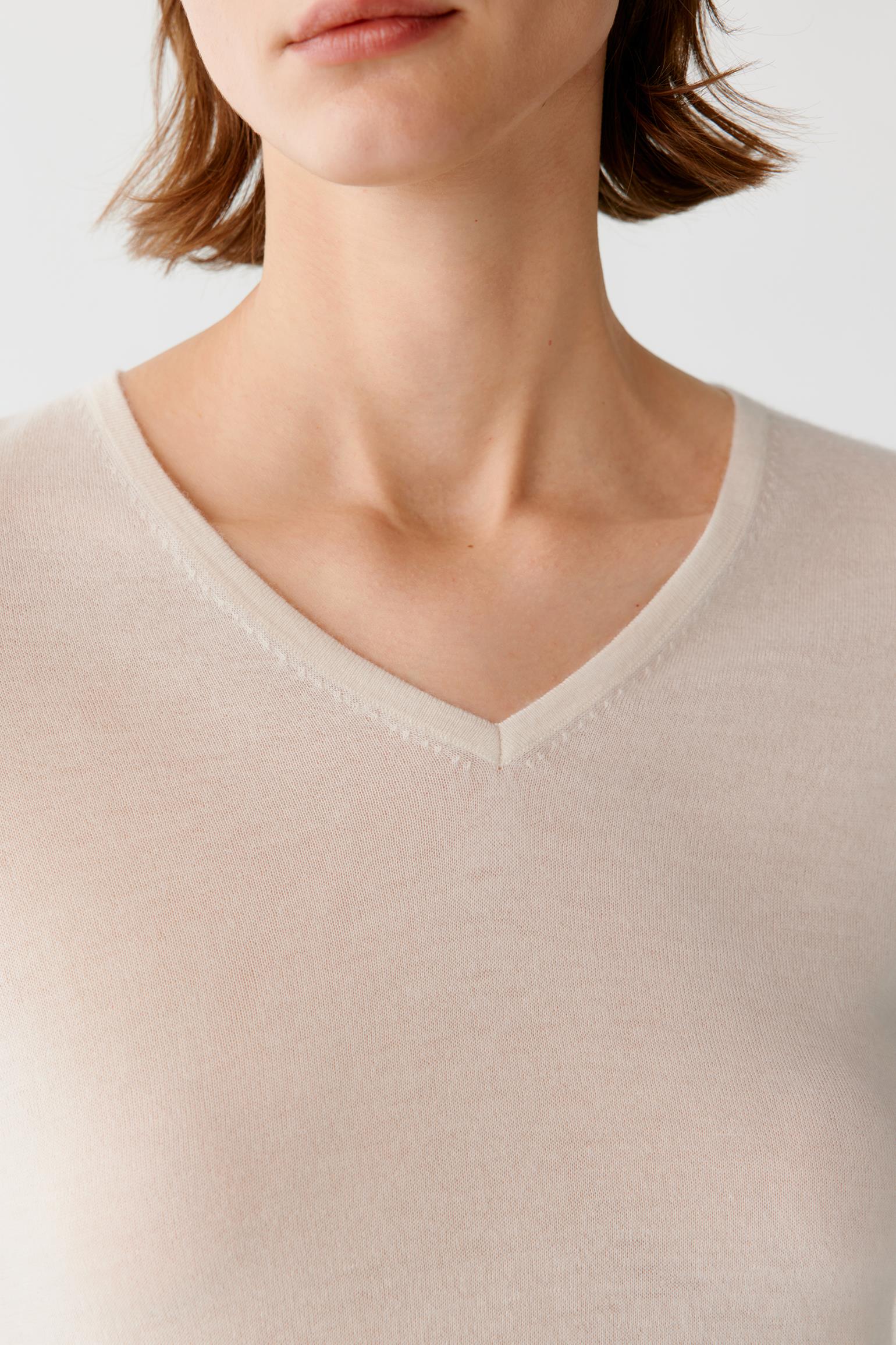 Close-up of a model wearing a light beige or cream V-neck knit top against a plain light gray background.