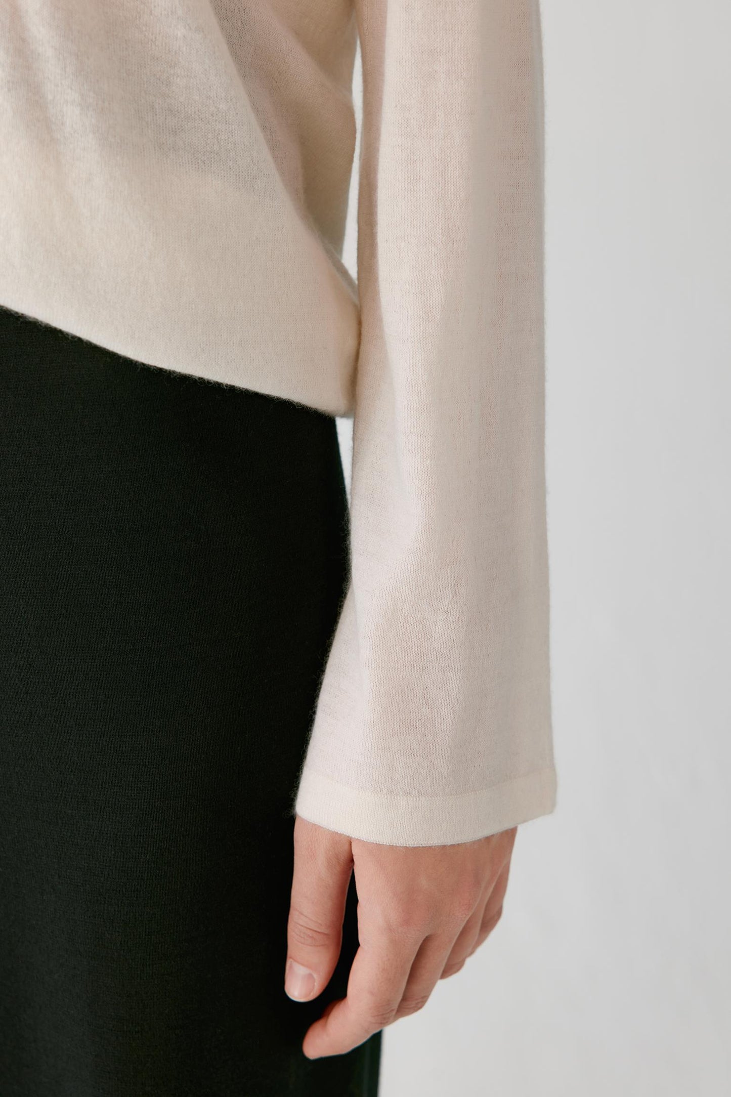 Close-up of a person wearing a light cream or off-white long-sleeve knit top with a black skirt or pants, against a plain light gray background.