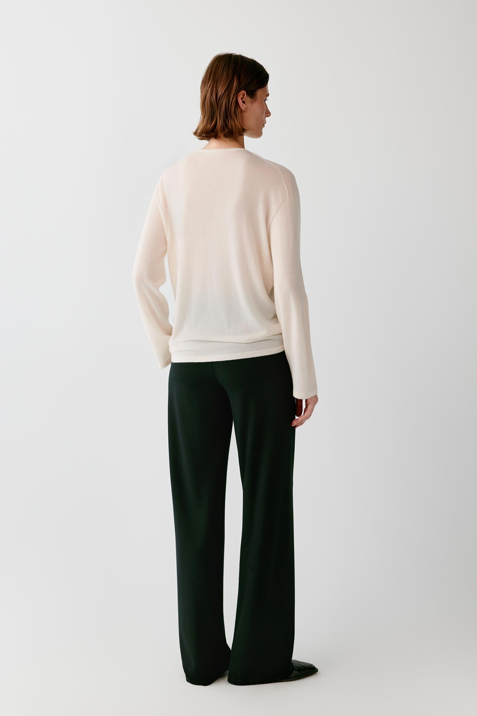 A model shown from the back wearing a cream-colored knit sweater and dark forest green wide-leg trousers, standing against a plain light gray studio background.