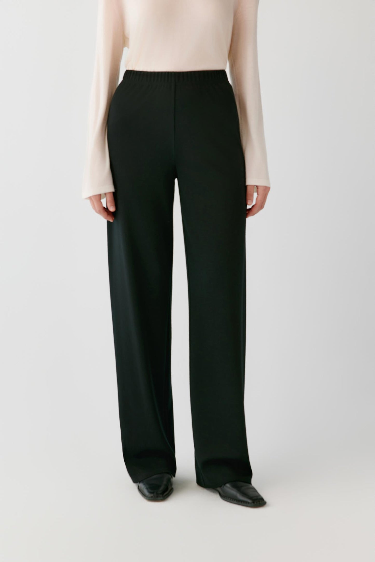 A model wearing black wide-leg trousers with a cream long-sleeve top and black pointed shoes, shown from the chest down against a light gray studio background.