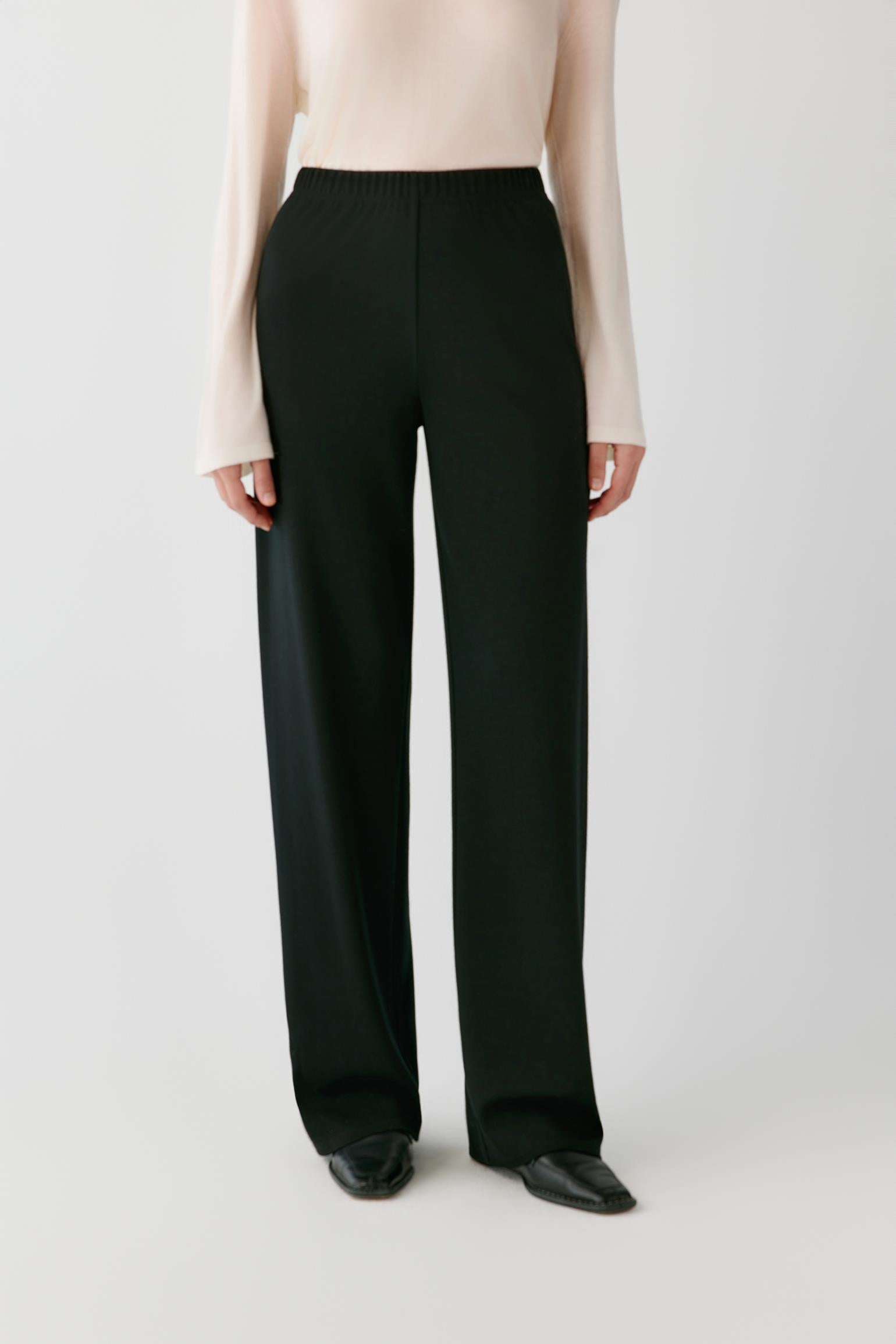 A model wearing black wide-leg trousers with a cream long-sleeve top and black pointed shoes, shown from the chest down against a light gray studio background.