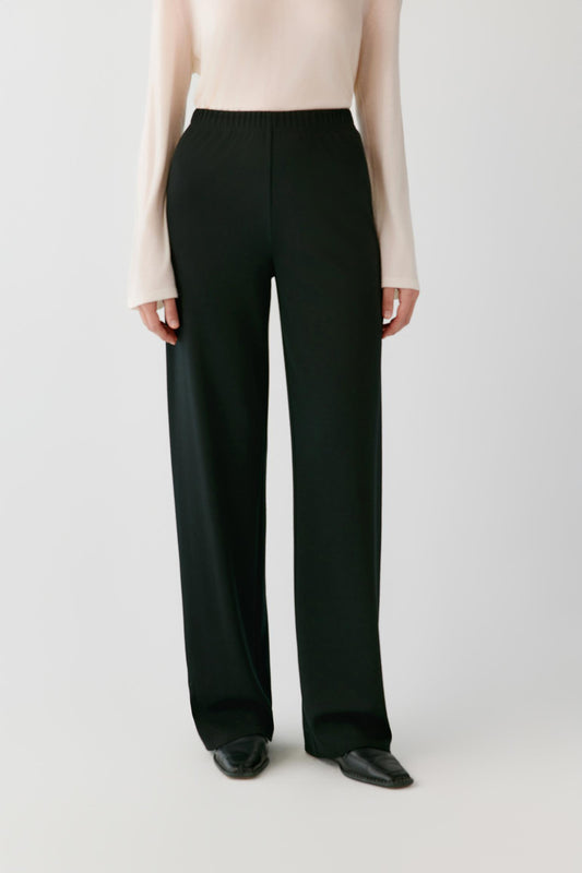 A model wearing black wide-leg trousers with a cream long-sleeve top and black pointed shoes, shown from the chest down against a light gray studio background.