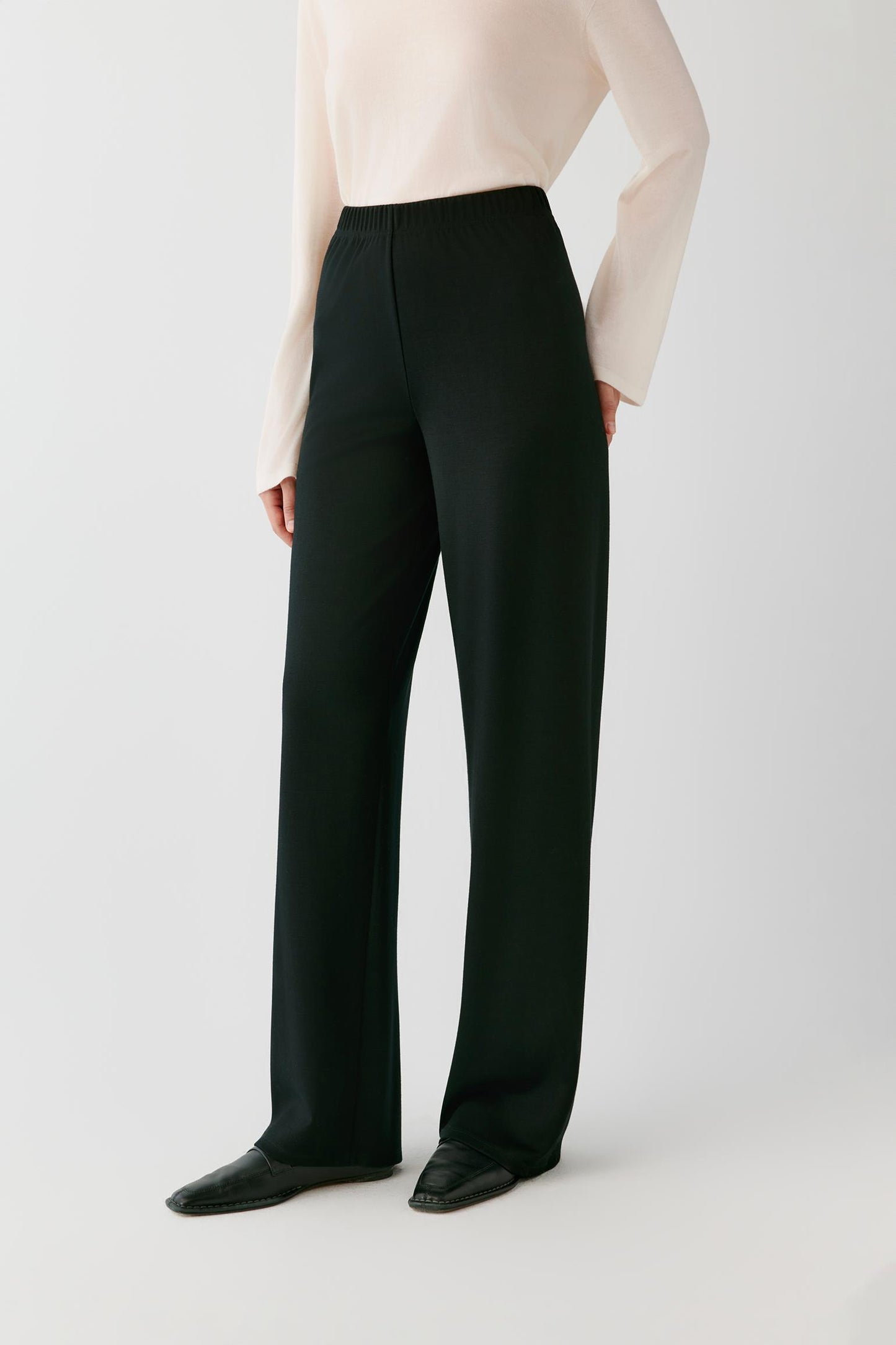 A model wearing black wide-leg trousers with a cream long-sleeve top and black pointed-toe flats, shown from the waist down against a light gray studio background.