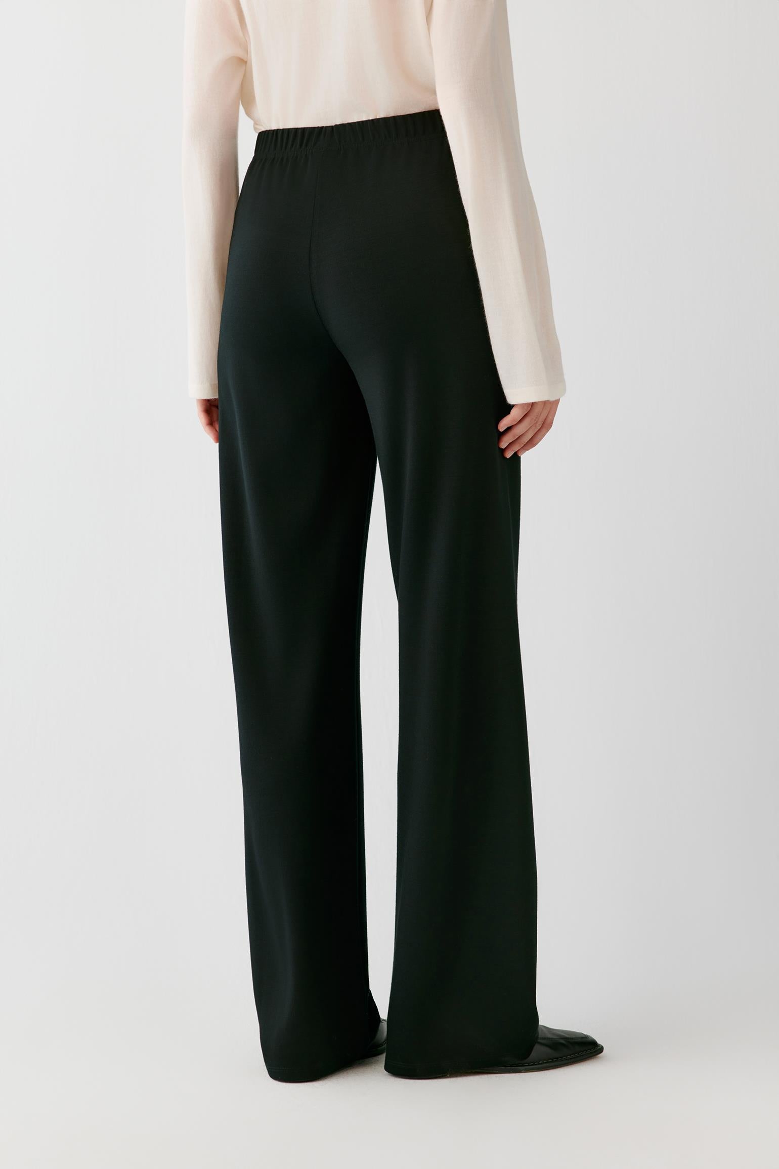 A person shown from the back wearing black wide-leg trousers and a light cream long-sleeve top against a plain light gray studio background.