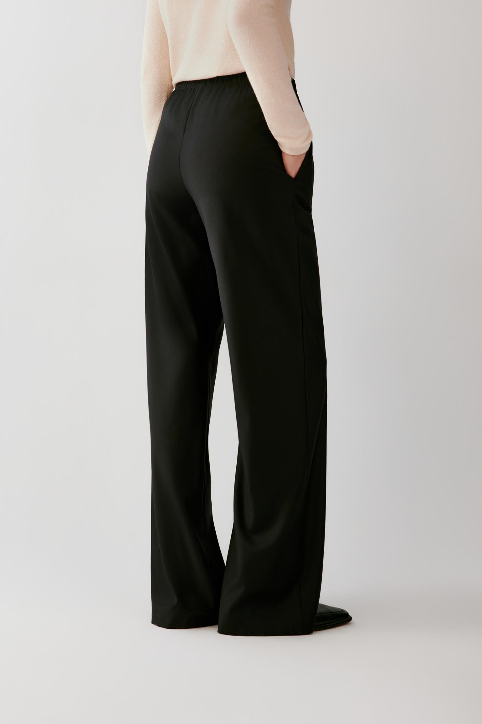 A model shown from the back wearing black wide-leg trousers and a light beige long-sleeve top, standing against a plain light gray studio background.