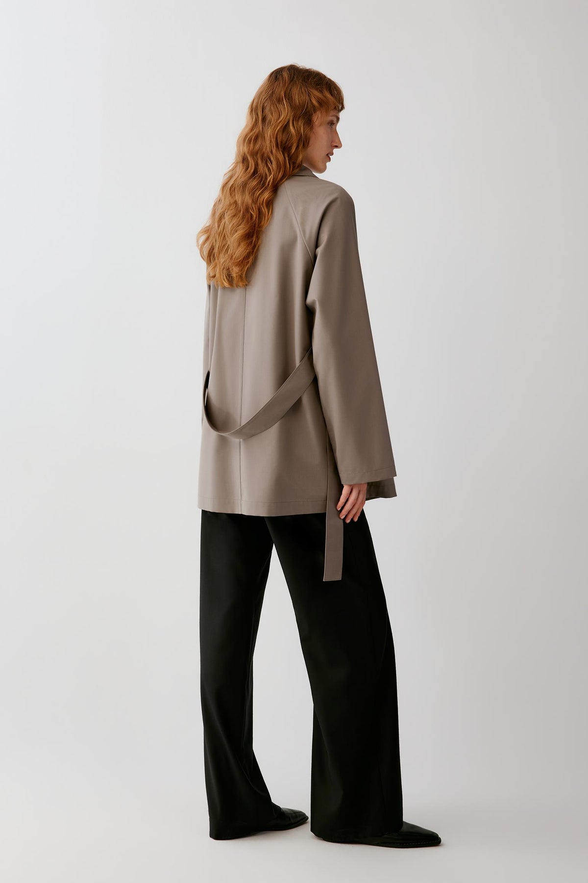 A model with long wavy red hair is shown from the back wearing a taupe oversized wrap-style blazer and black wide-leg trousers against a light gray studio background.
