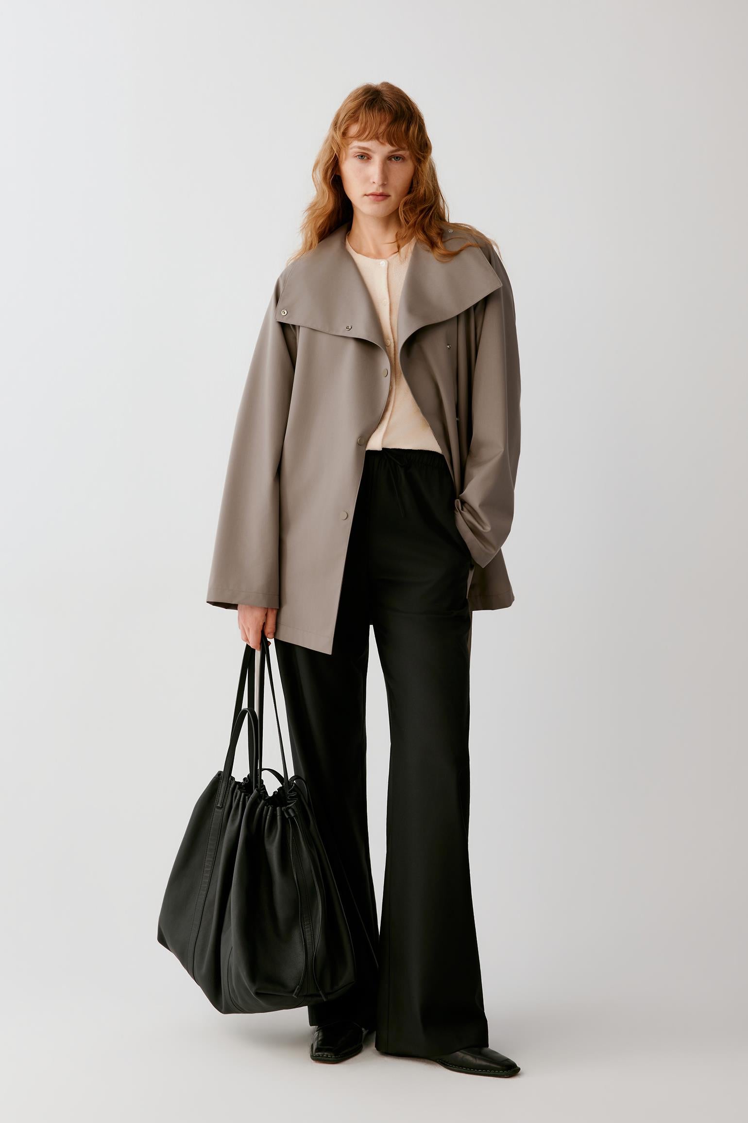 A model stands against a light gray studio background wearing a taupe oversized wrap-style coat over a cream top and black wide-leg trousers, while holding a large black leather tote bag.