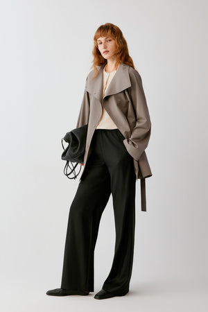 A woman modeling a taupe-brown wrap coat over a cream top and black wide-leg trousers, holding a black handbag against a light gray studio background.