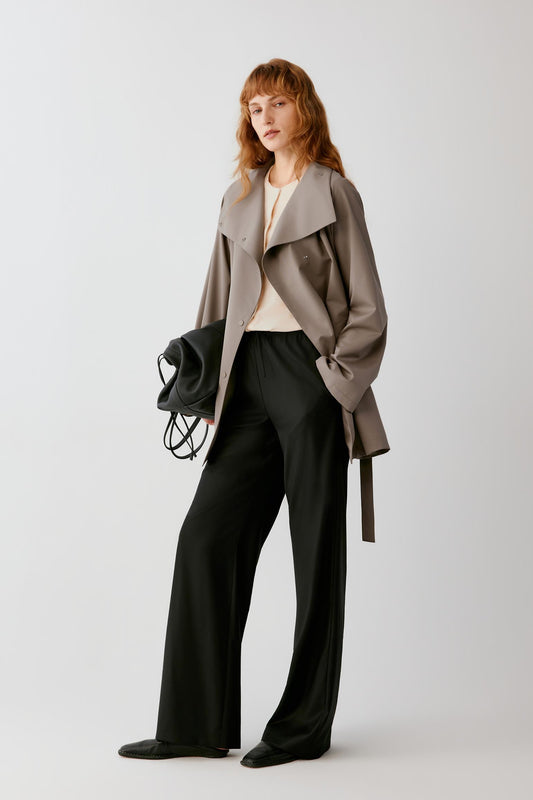 A woman modeling a taupe-brown wrap coat over a cream top and black wide-leg trousers, holding a black handbag against a light gray studio background.