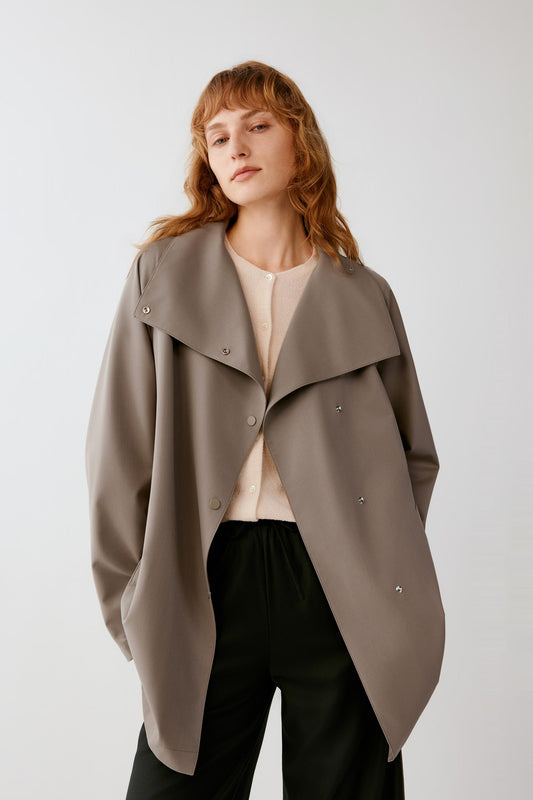 A woman wearing a taupe oversized wrap-style coat over a cream top and black pants, photographed against a light gray studio background.