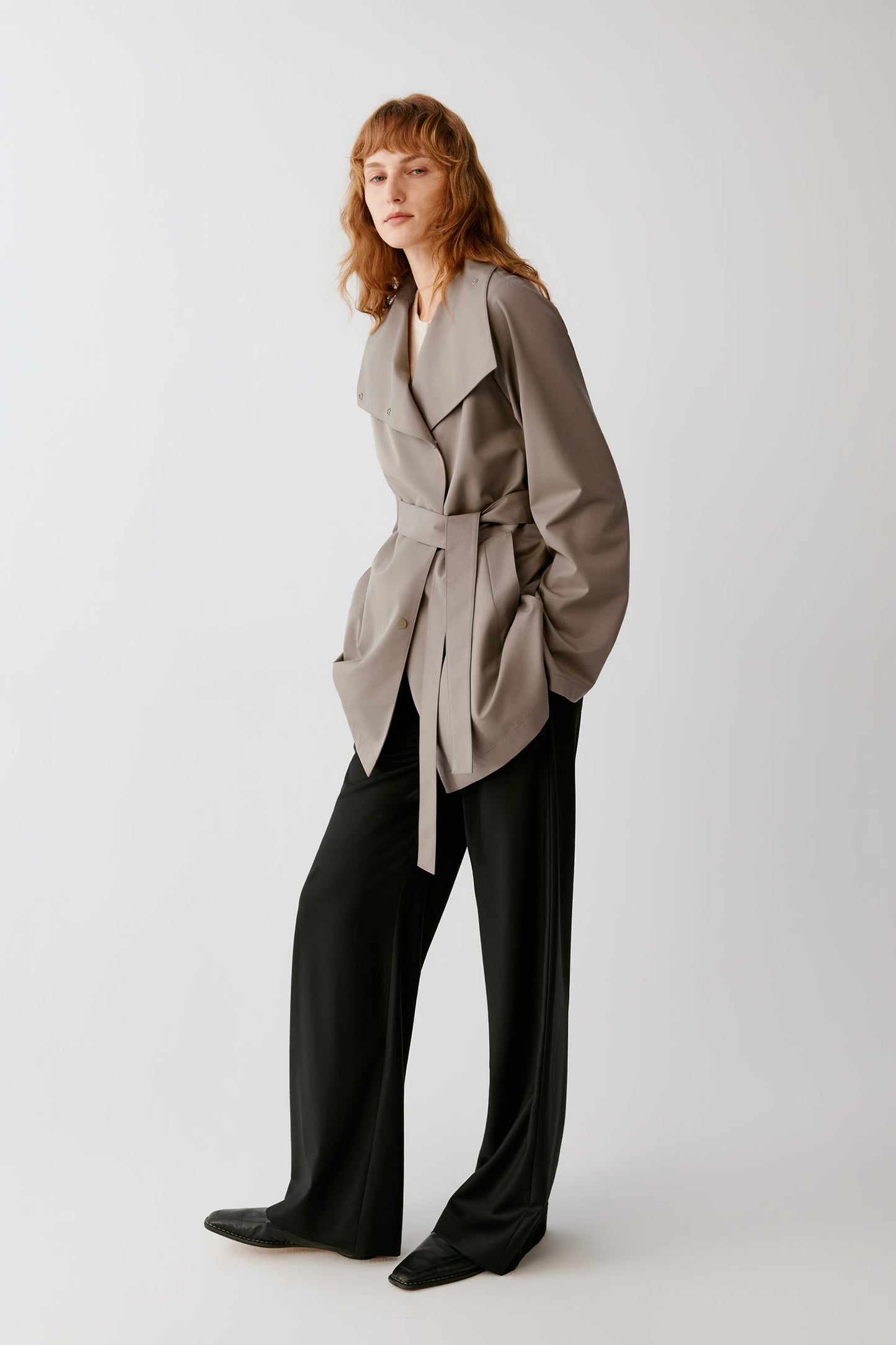 A woman modeling a taupe wrap-style belted jacket over a black wide-leg jumpsuit, standing against a light gray studio background.