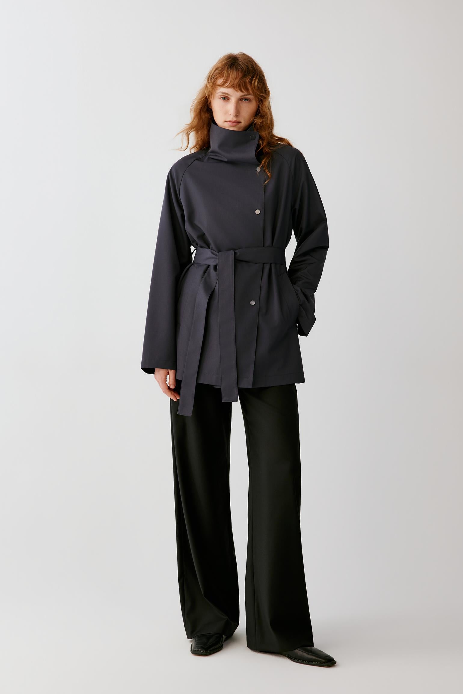 Front view of a woman wearing a deep charcoal grey wool trench coat, highlighting the long silhouette, high stand collar and belted waist.