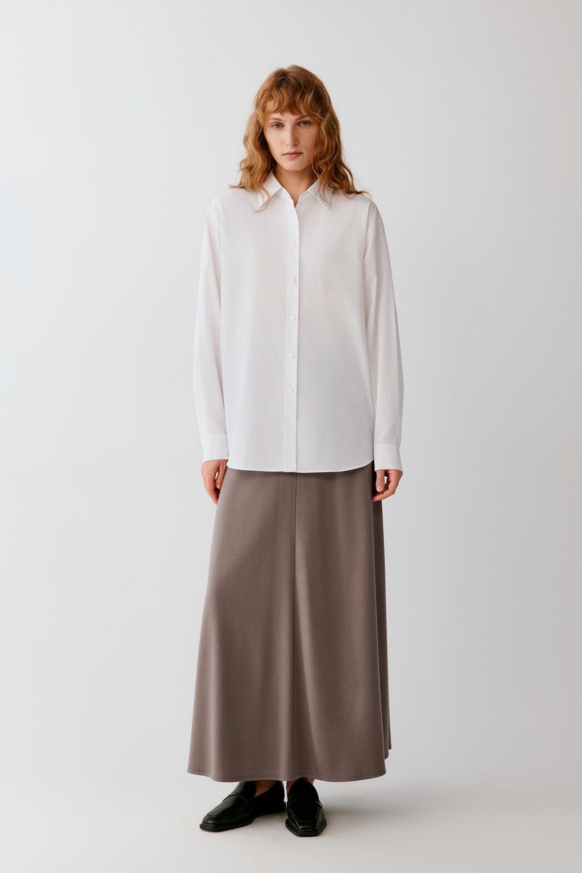 A woman stands against a plain light gray studio background wearing a loose white long-sleeve button-up shirt and a long taupe-brown skirt, with black shoes.