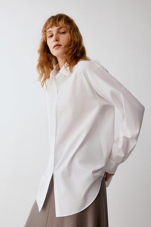 A woman with wavy light brown hair wearing an oversized white button-up shirt and a taupe skirt, posing against a light gray studio background.