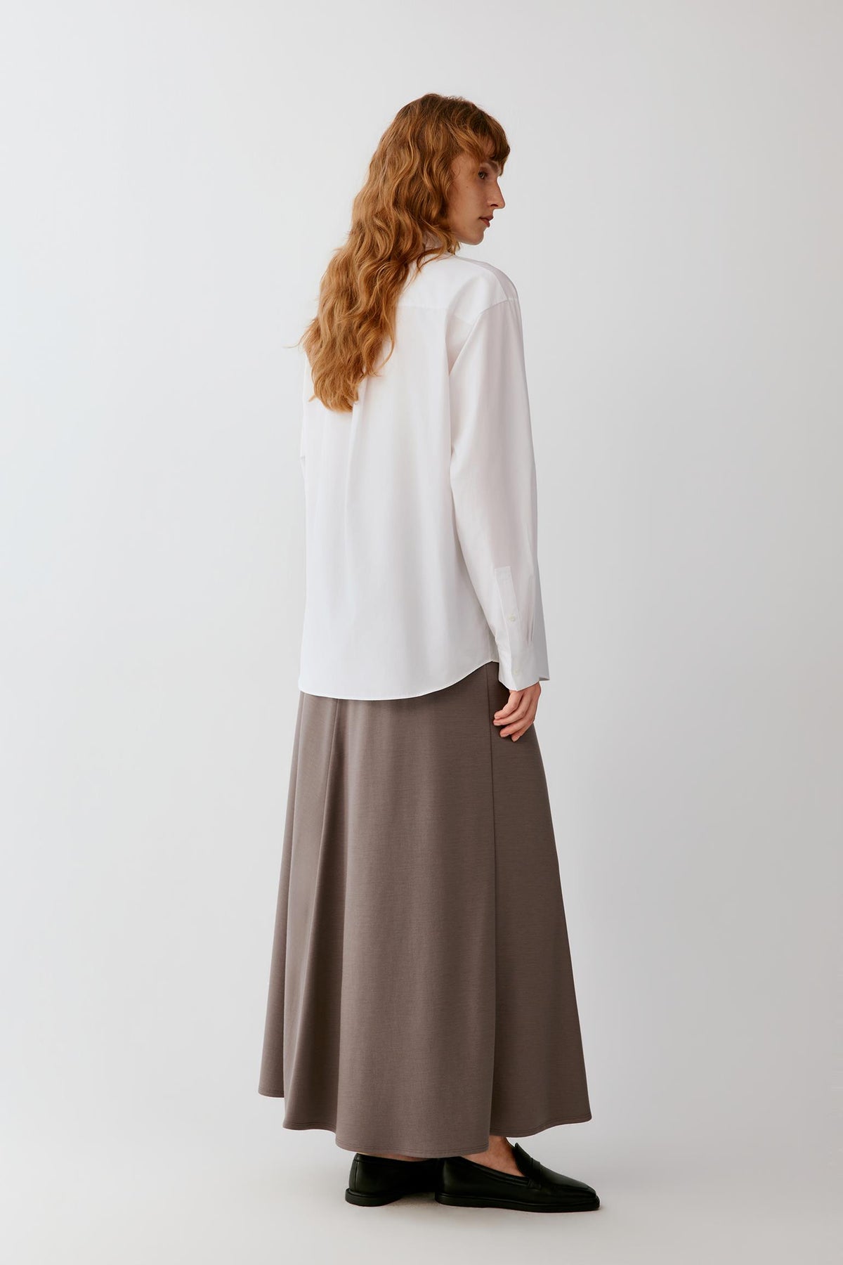 A woman with long wavy light brown hair is shown from the back wearing a white long-sleeve blouse and a high-waisted, flowing taupe-brown skirt. She is standing against a plain, light gray studio background.
