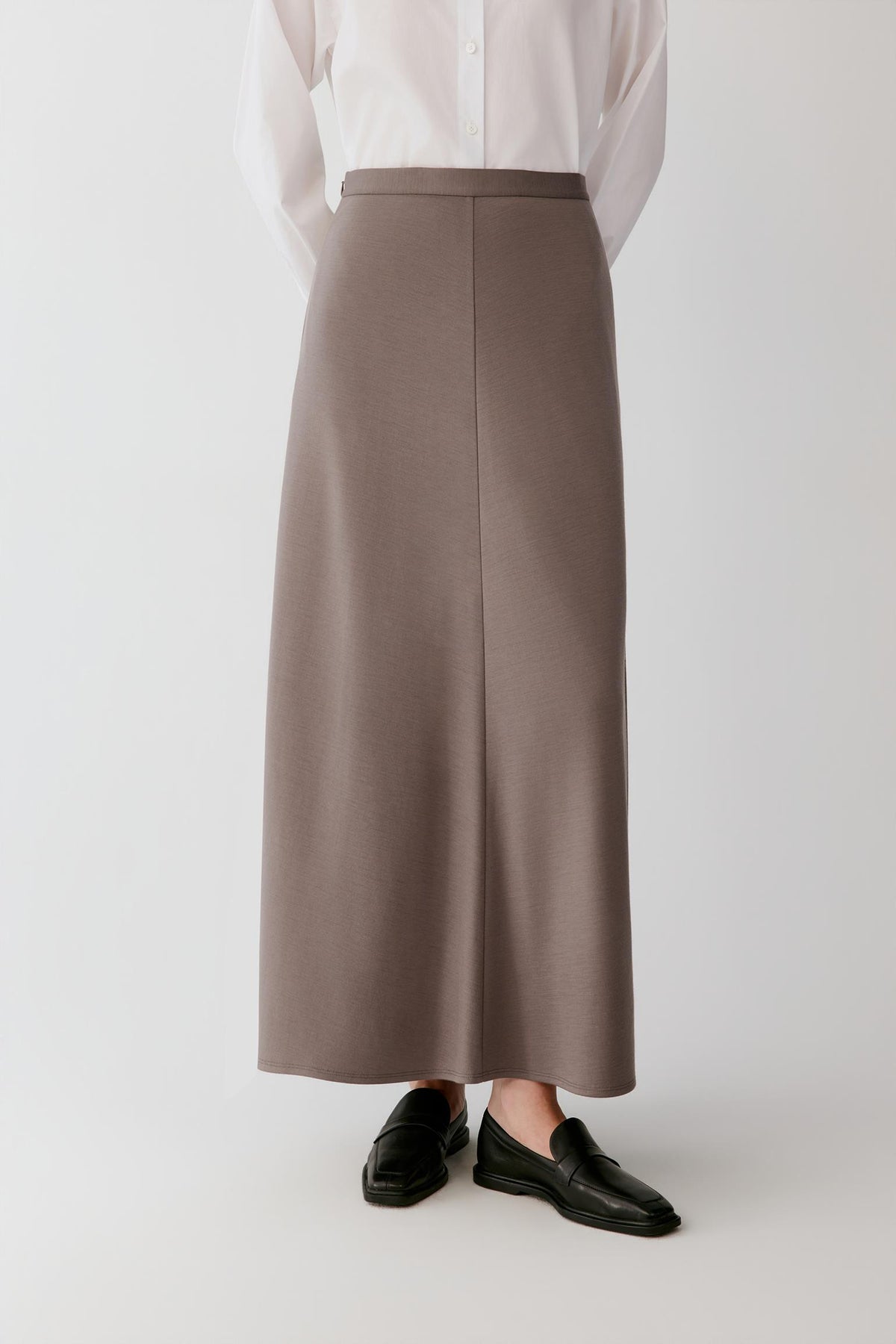 A person wearing a long taupe-brown midi skirt with a front center seam, paired with a white blouse and black loafers, shown from the waist down against a light gray studio background.