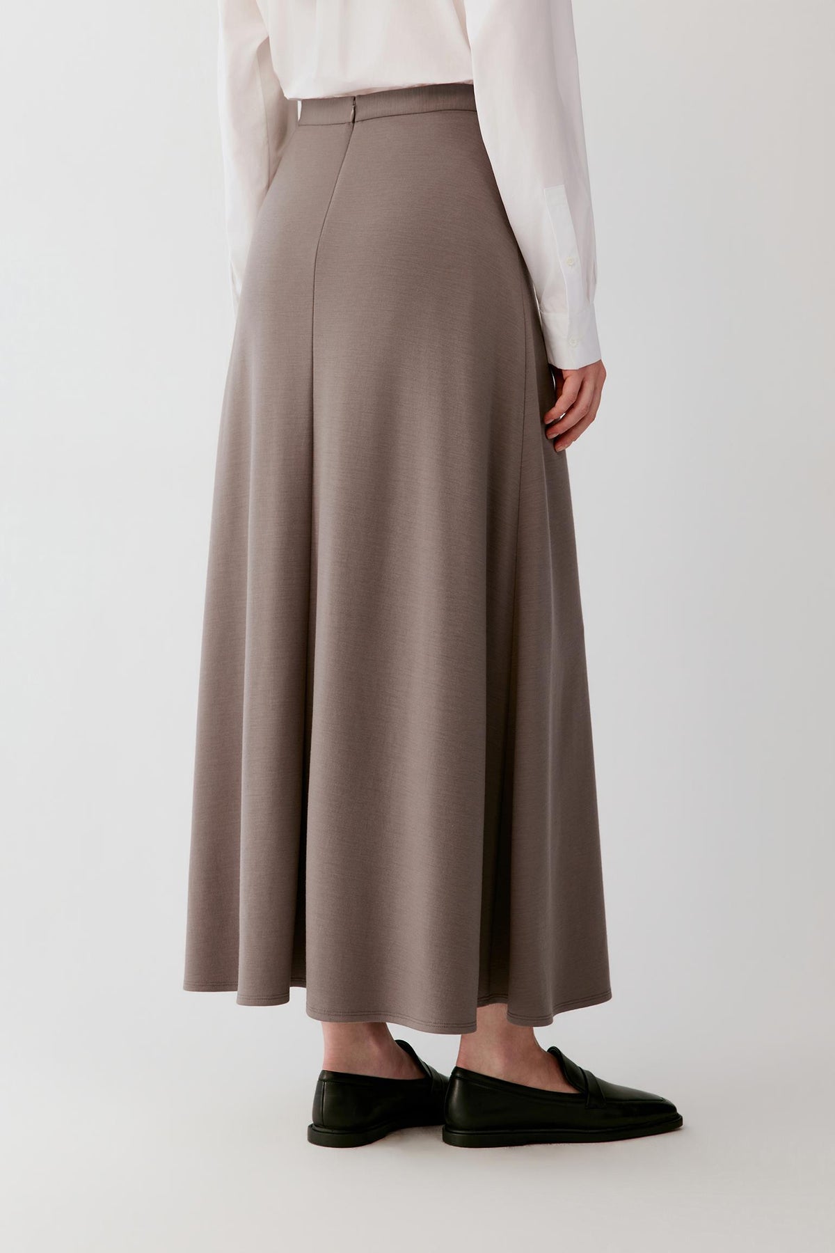 A person shown from the waist down wearing a long, high-waisted taupe skirt with a subtle sheen, paired with a white long-sleeve blouse and black loafers, against a plain light gray studio background.