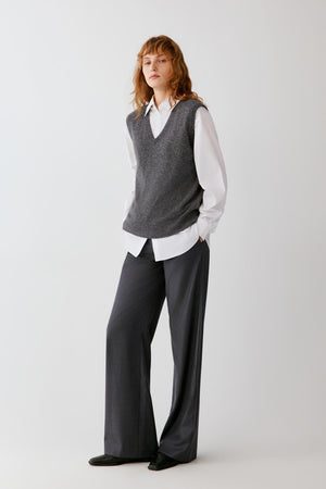 A woman stands against a light gray studio background wearing a gray knit sweater vest over a white collared shirt with dark charcoal wide-leg trousers and black shoes.