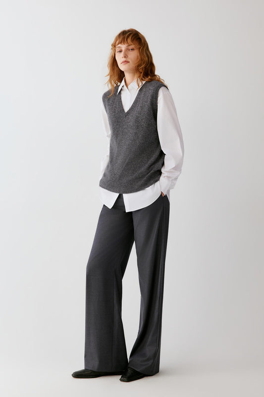 A woman stands against a light gray studio background wearing a gray knit sweater vest over a white collared shirt with dark charcoal wide-leg trousers and black shoes.