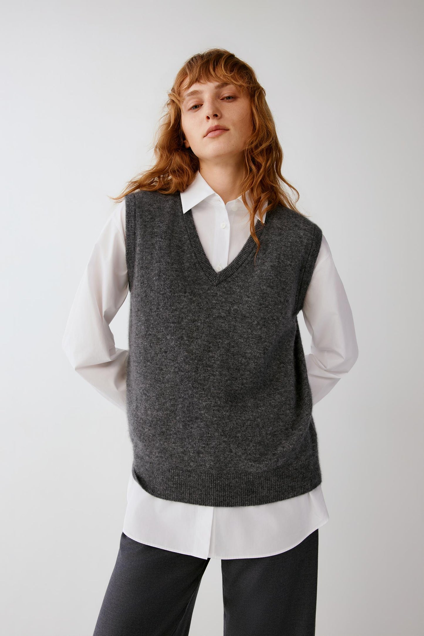 A model wearing a dark gray knit sweater vest over a white collared shirt and dark trousers, photographed against a plain light gray studio background.