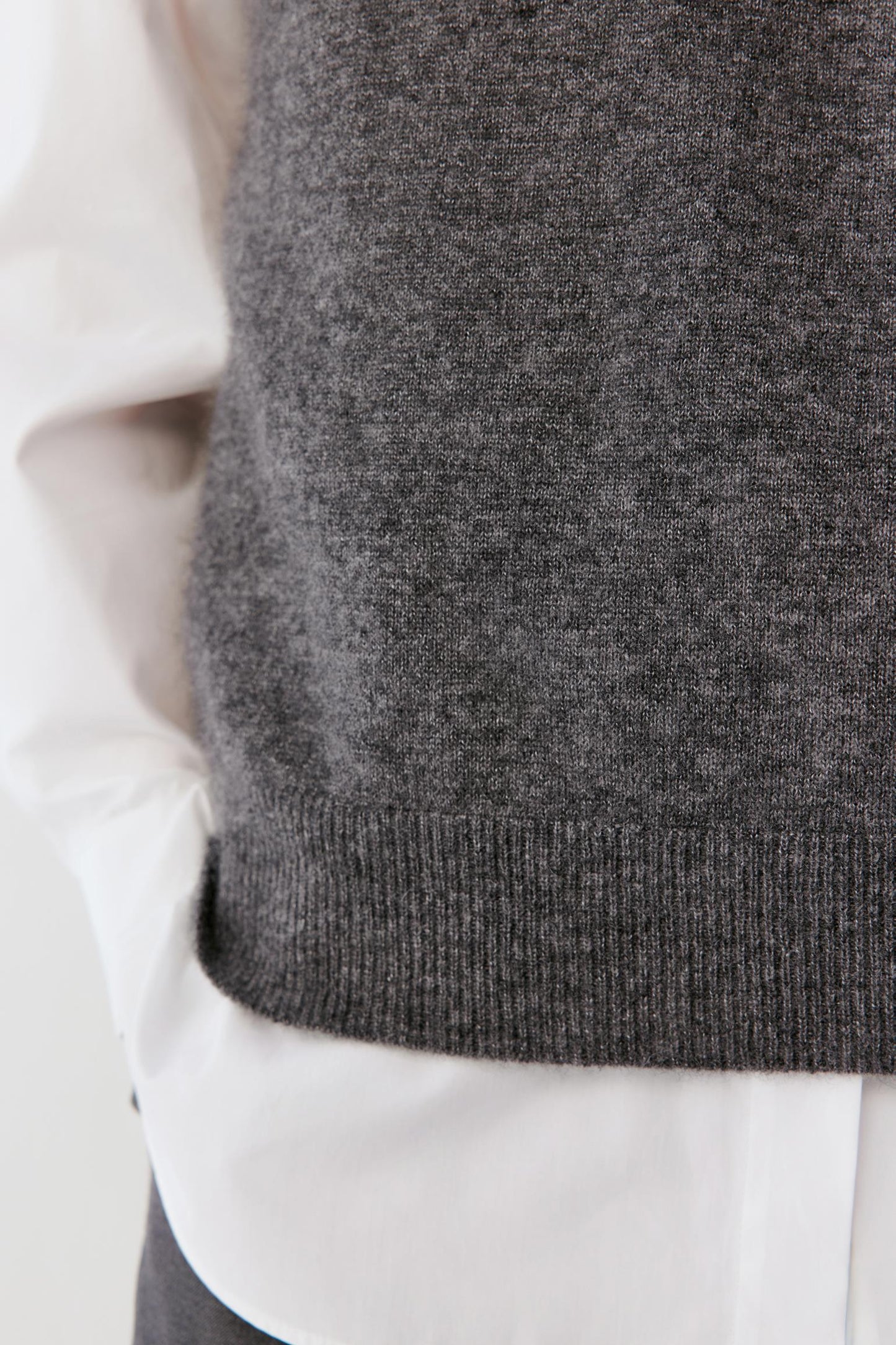 Close-up of a dark gray knit sweater vest layered over a white collared shirt, showing the ribbed hem and textured fabric.