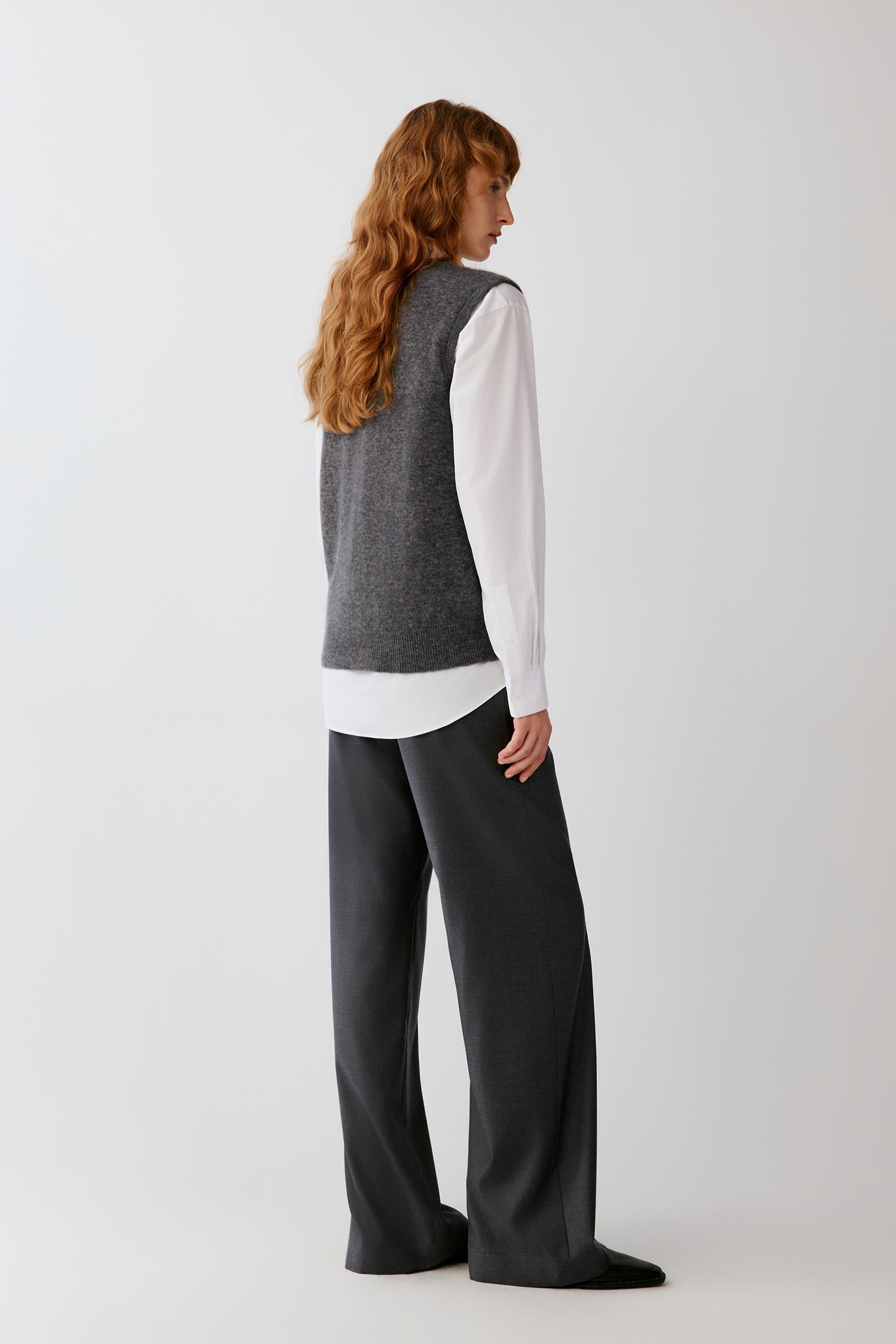 A woman with long wavy red hair is shown from the back wearing a gray sleeveless knit vest over a white long-sleeve top and dark charcoal wide-leg trousers, photographed against a light gray studio background.