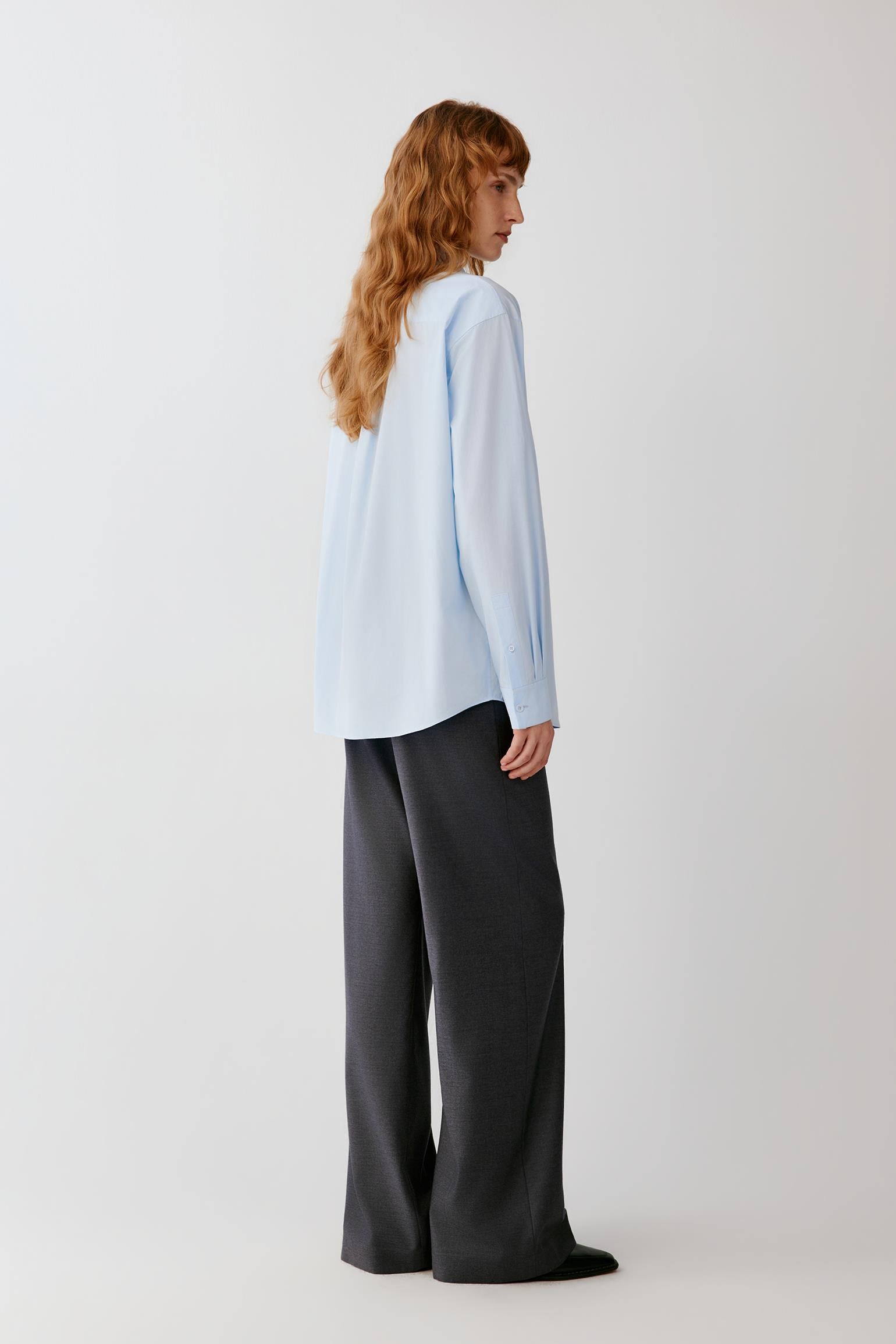 A woman with long wavy light brown hair is shown from the back wearing a light blue long-sleeve blouse and dark charcoal gray wide-leg trousers, standing against a plain light gray studio background.