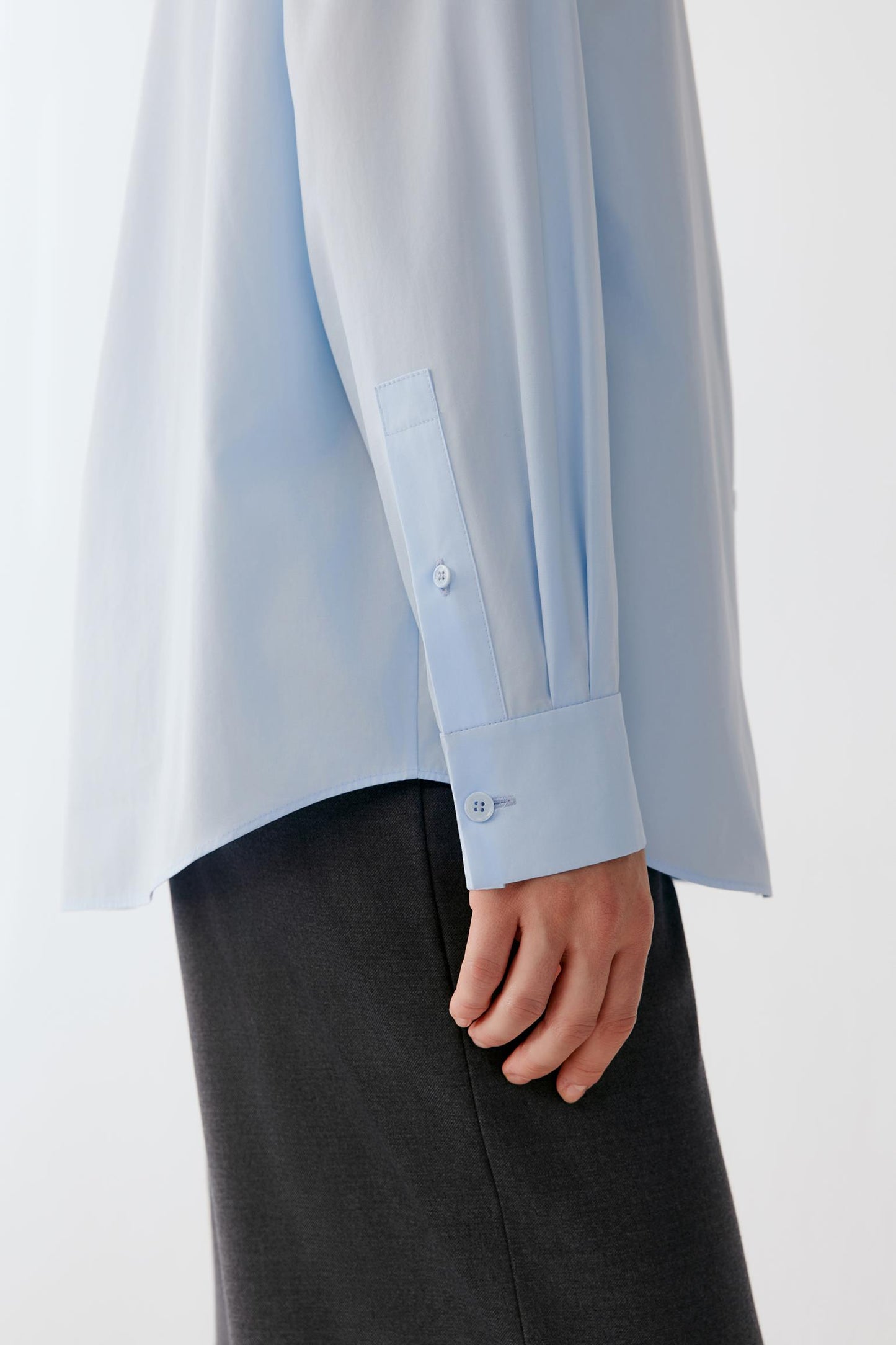 A close-up of a person wearing a light blue long-sleeve button-up shirt with a chest pocket and dark charcoal gray trousers, shown against a plain light background.