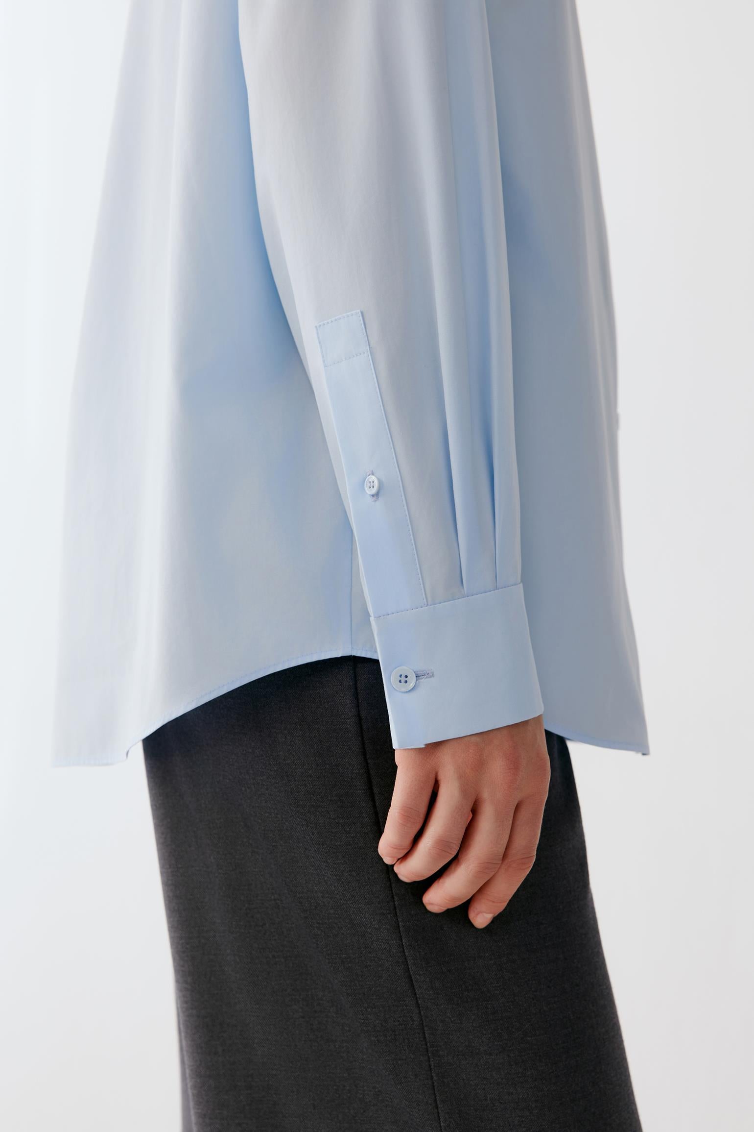 A close-up of a person wearing a light blue long-sleeve button-up shirt with a chest pocket and dark charcoal gray trousers, shown against a plain light background.