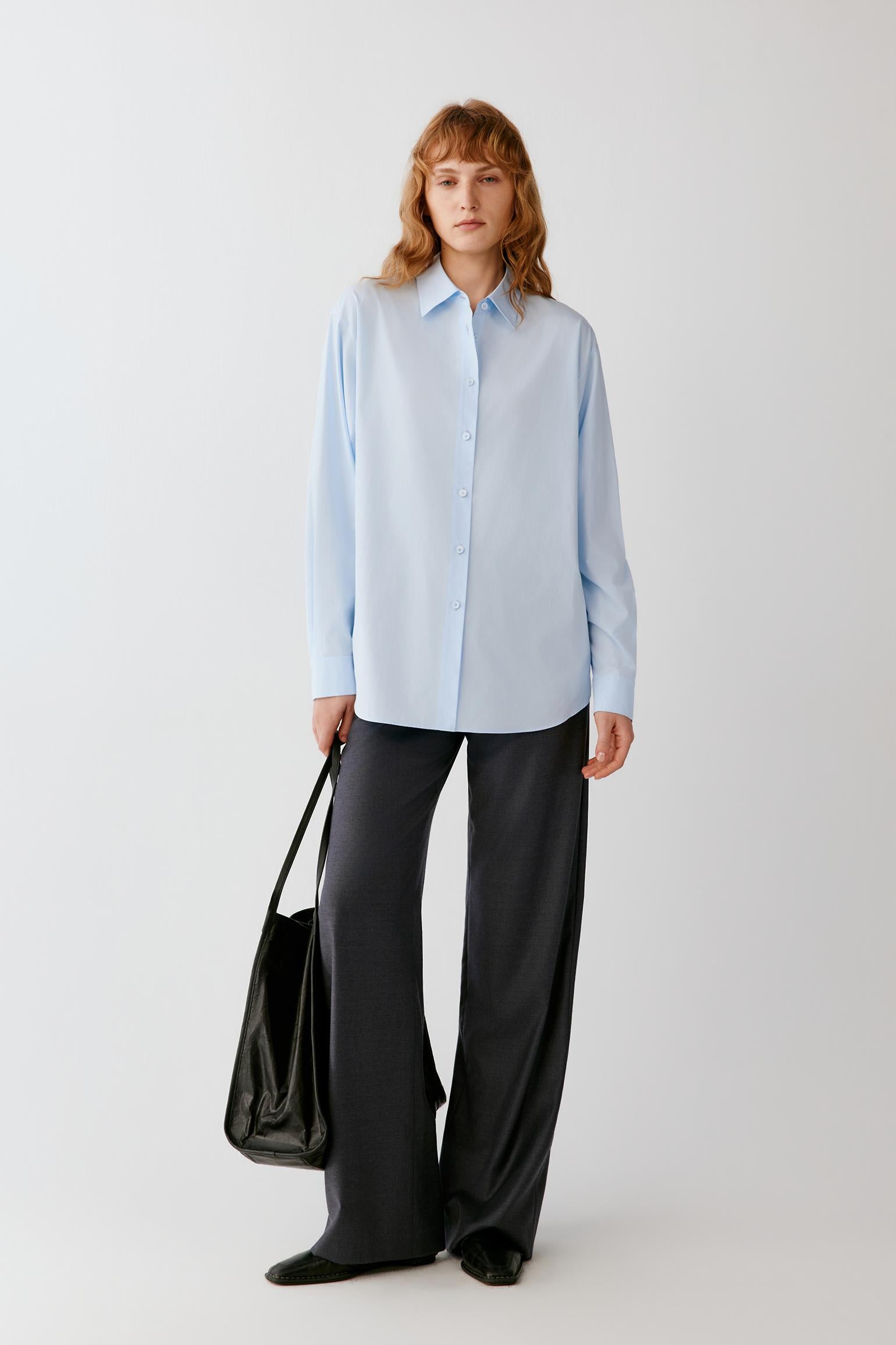A woman stands against a light gray studio background wearing a light blue long-sleeve button-up shirt and dark wide-leg pants, while holding a black shoulder bag.