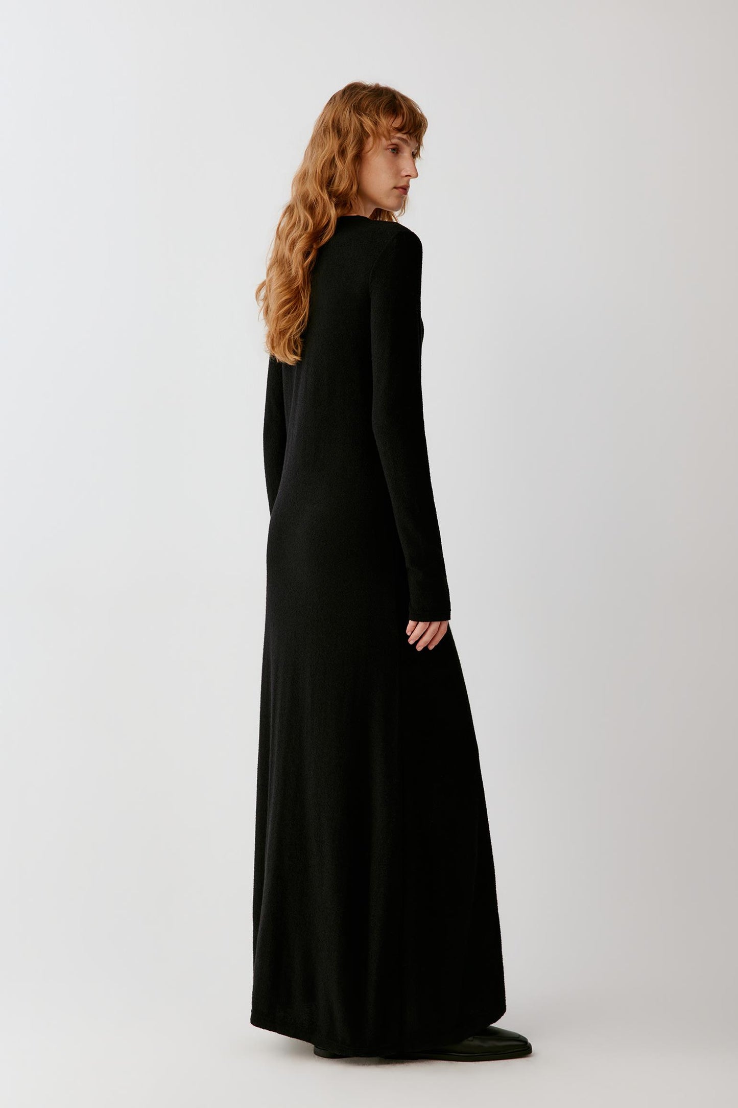 Woman wearing a long black fitted dress with long sleeves and black loafers.