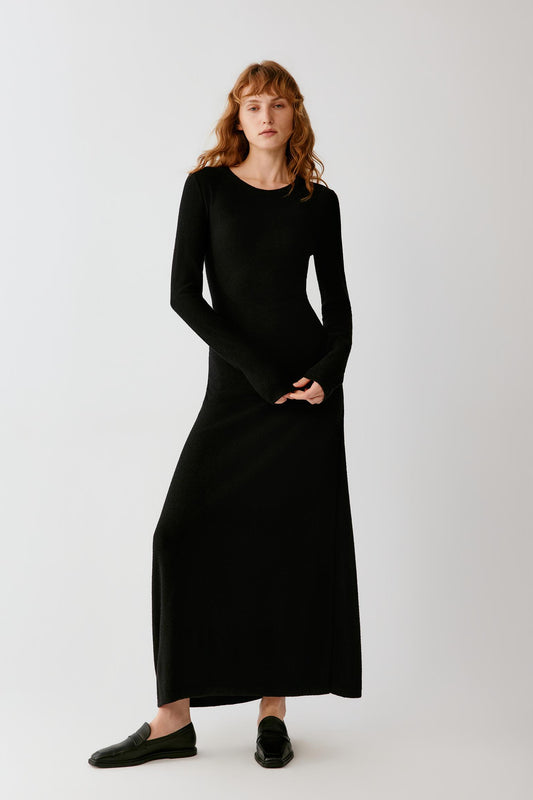 Woman wearing a long black fitted dress with long sleeves and black loafers.