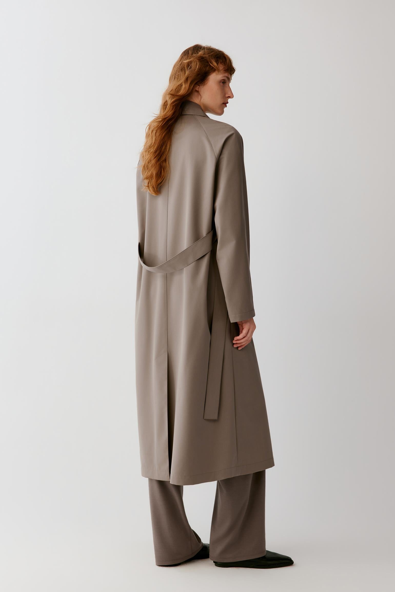 A model shown from the back wearing a long taupe-brown belted coat, wide-leg trousers, and black shoes against a plain light gray studio background.