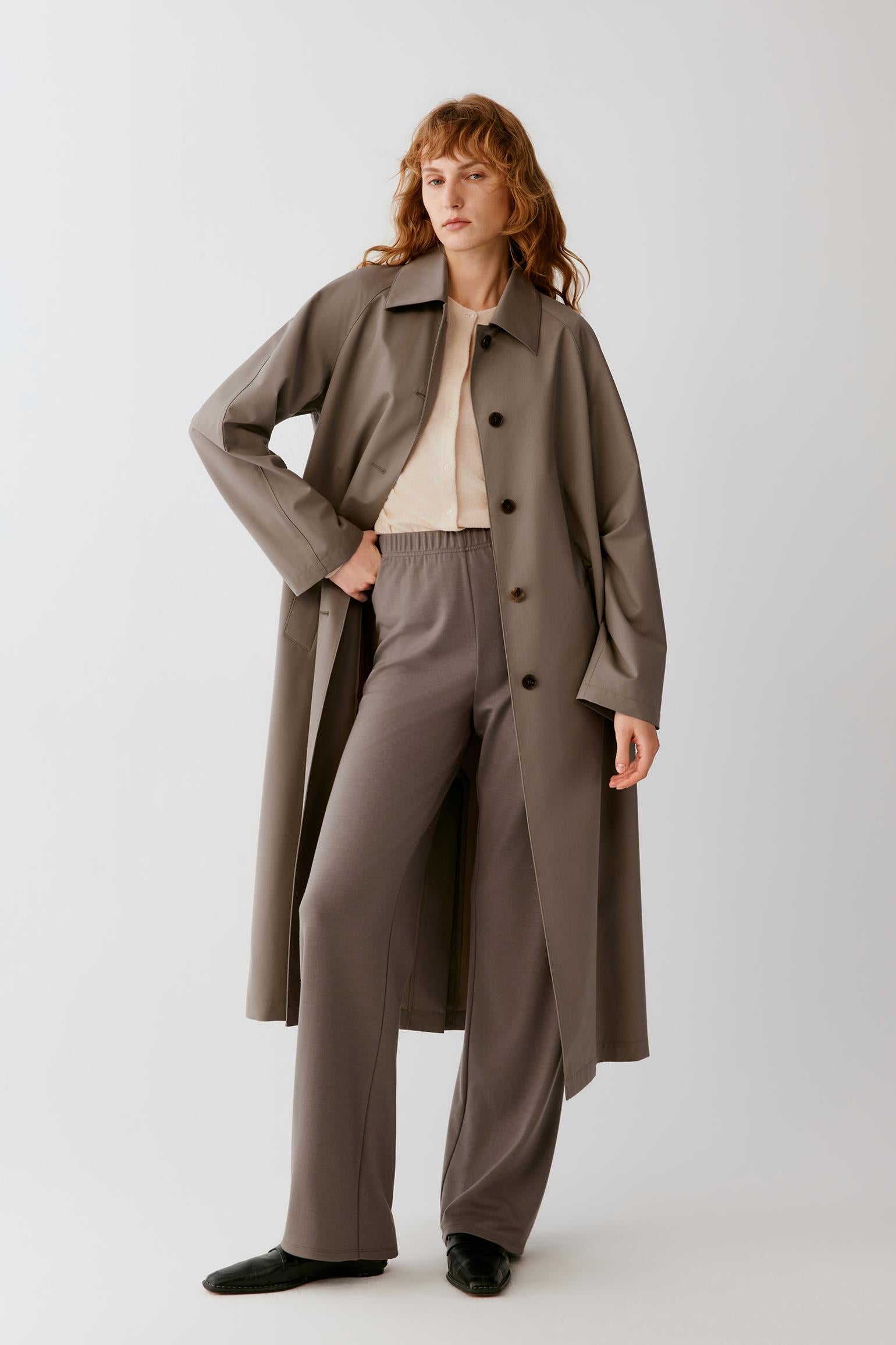 A woman modeling a long taupe-brown trench coat over a cream top and matching wide-leg trousers, photographed against a light gray studio background.