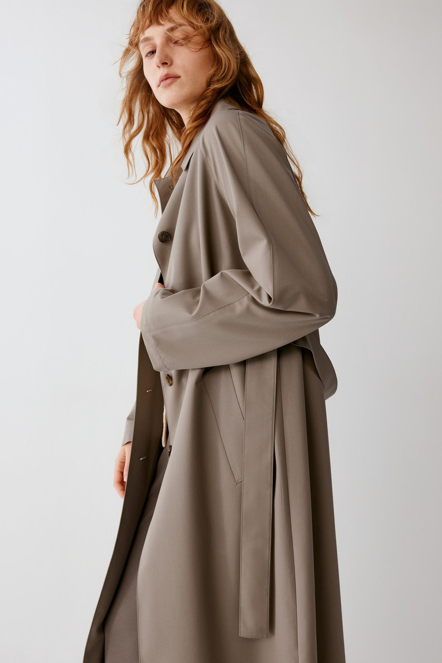 A woman wearing a long taupe-brown trench coat with a belted waist, shown in a side profile against a plain light gray studio background.